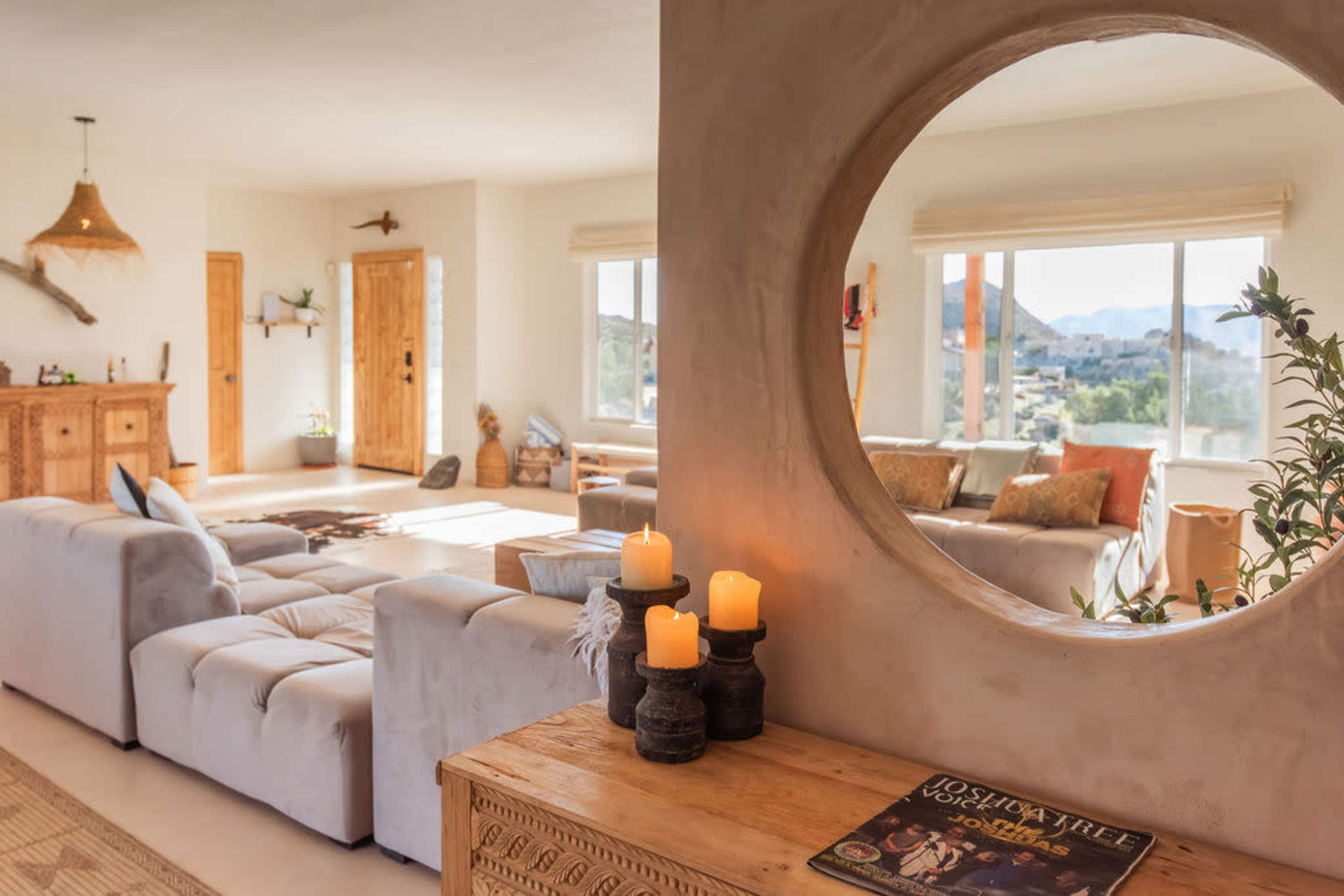 The image shows a bright, modern living space featuring soft furniture, a circular mirror, and decorative items, with large windows revealing a desert landscape outside.