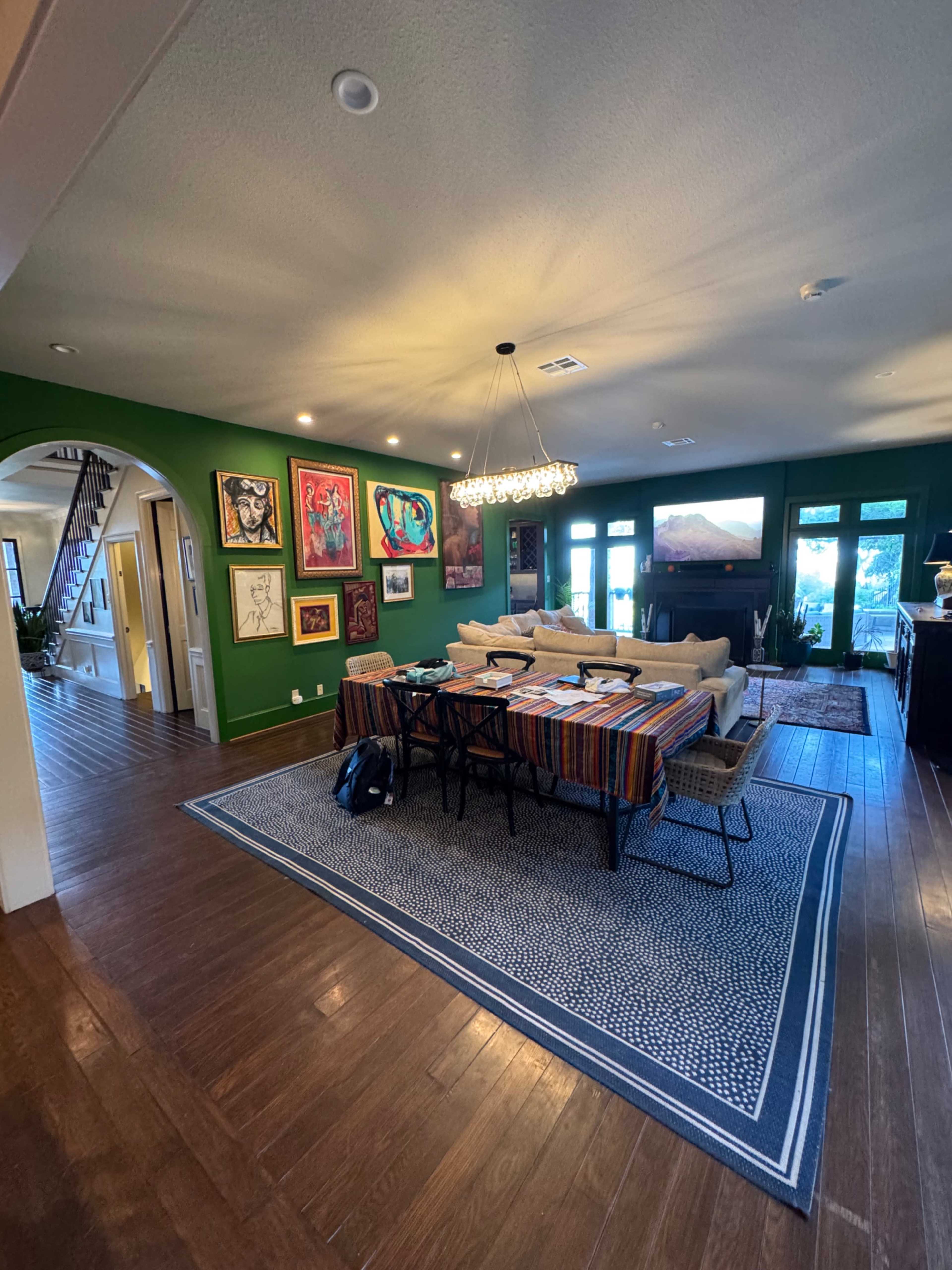 A spacious living area features a green accent wall adorned with artwork, a large sofa, a dining table, and ample natural light from surrounding windows.