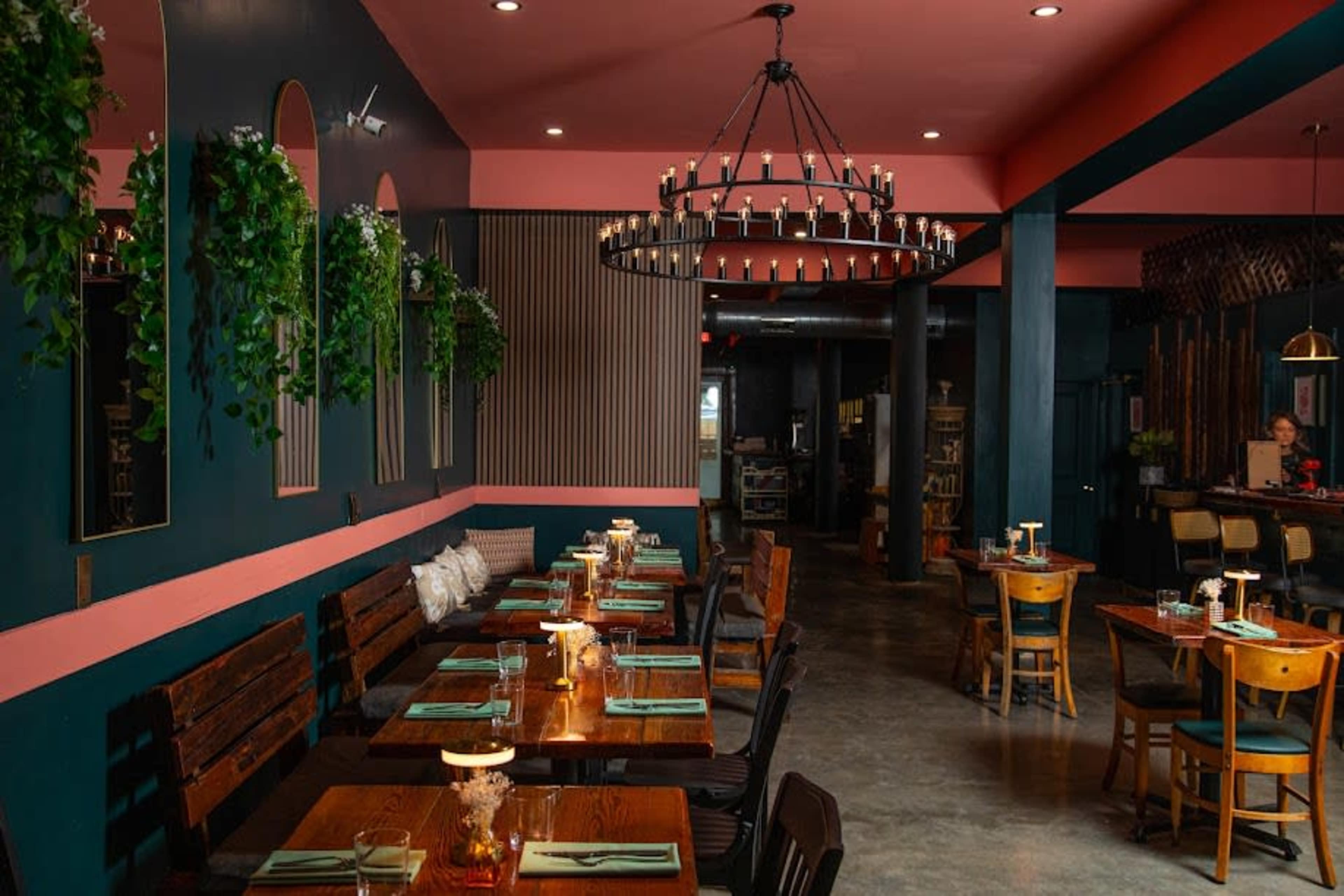 The image shows a modern restaurant interior featuring wooden tables, chairs, and decorative plants, with a large chandelier hanging from the ceiling.
