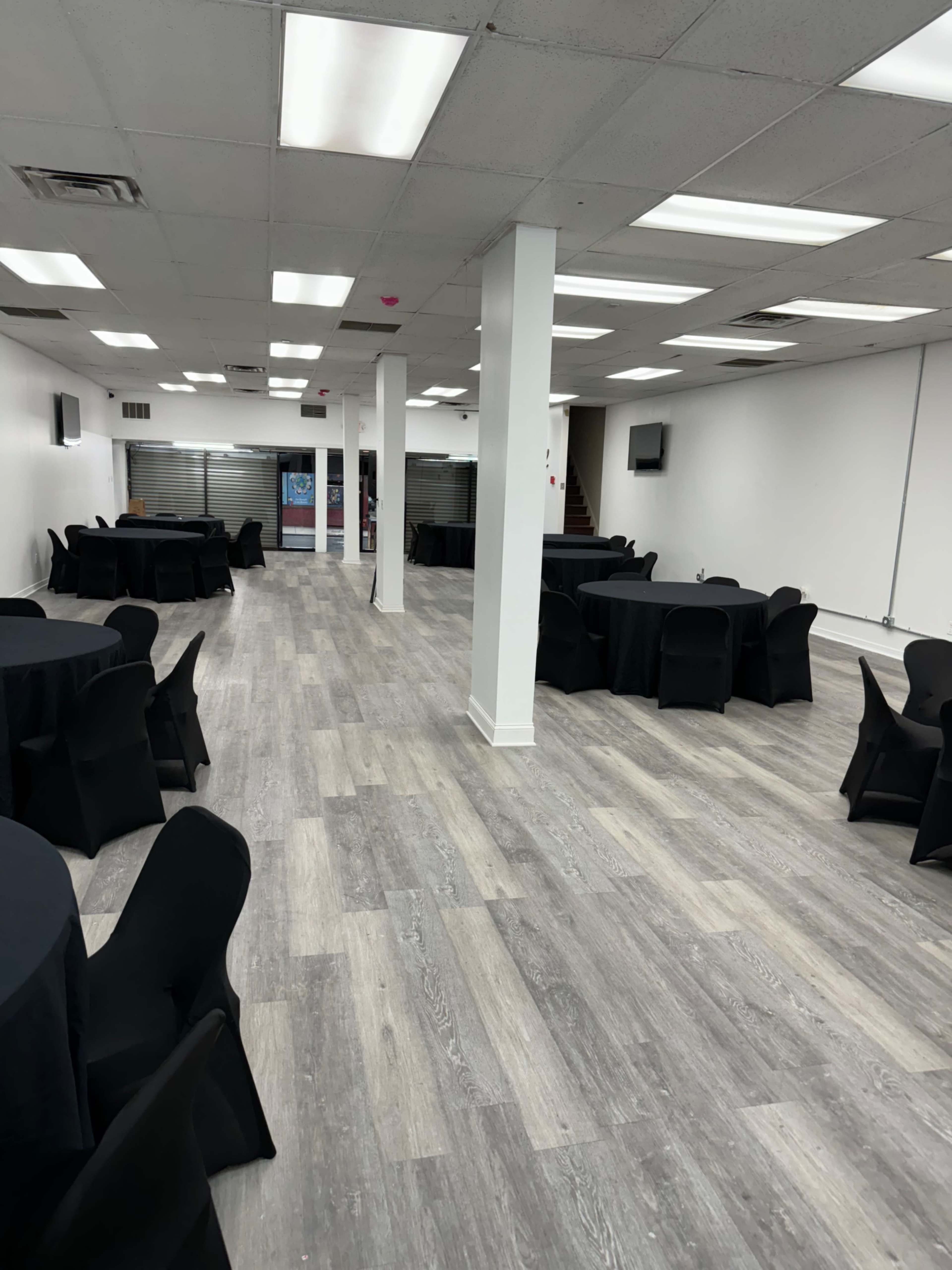 A spacious event room with black tablecloths covering round tables arranged throughout, and a light-colored wooden floor.