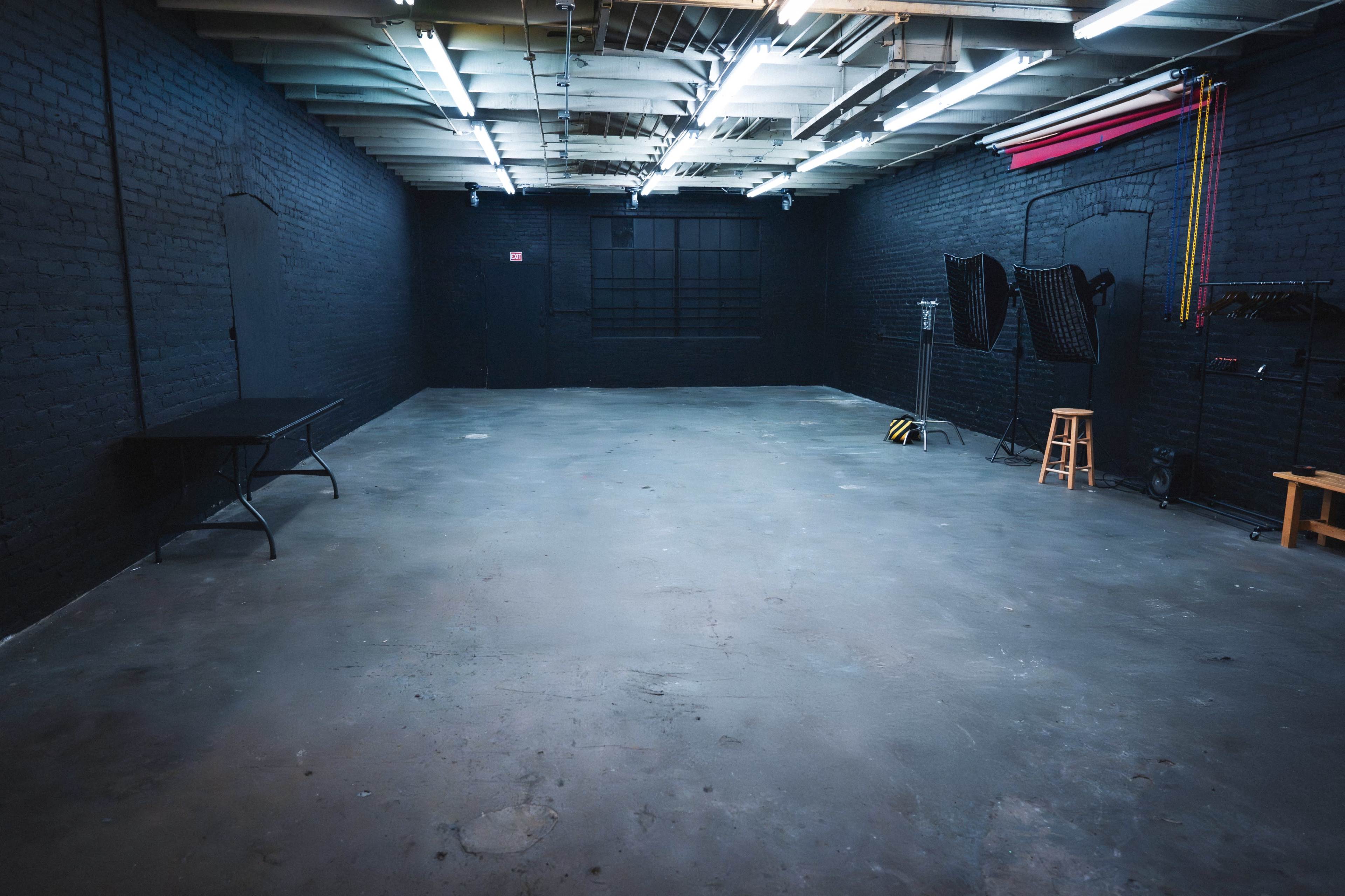 The image shows a large, empty studio space with exposed beams, a concrete floor, and minimal lighting fixtures along the ceiling.