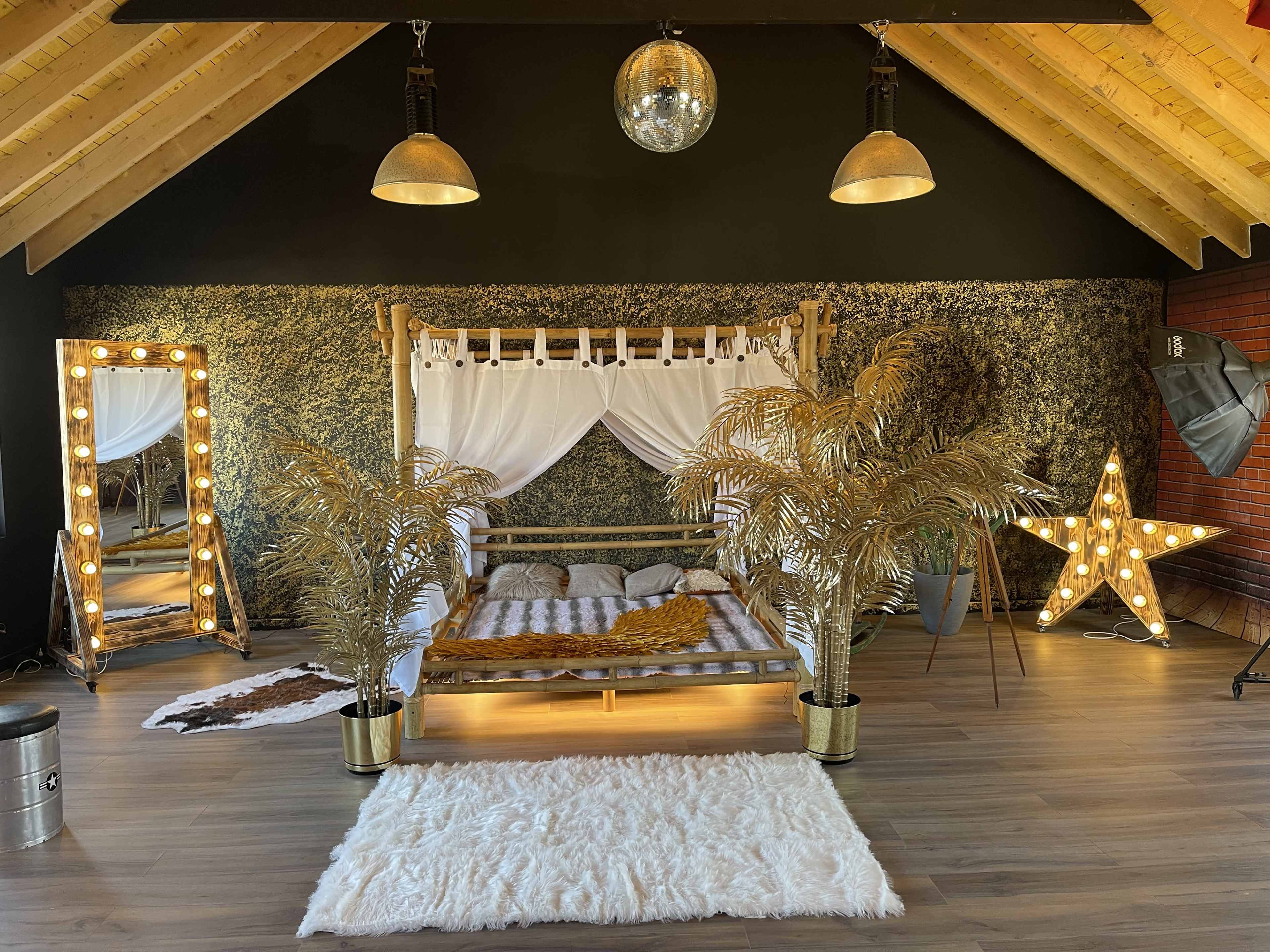 The image shows a stylish bedroom with a wooden bed frame, a large mirror surrounded by lights, decorative plants, and a star-shaped light fixture against a black wall and wooden ceiling.