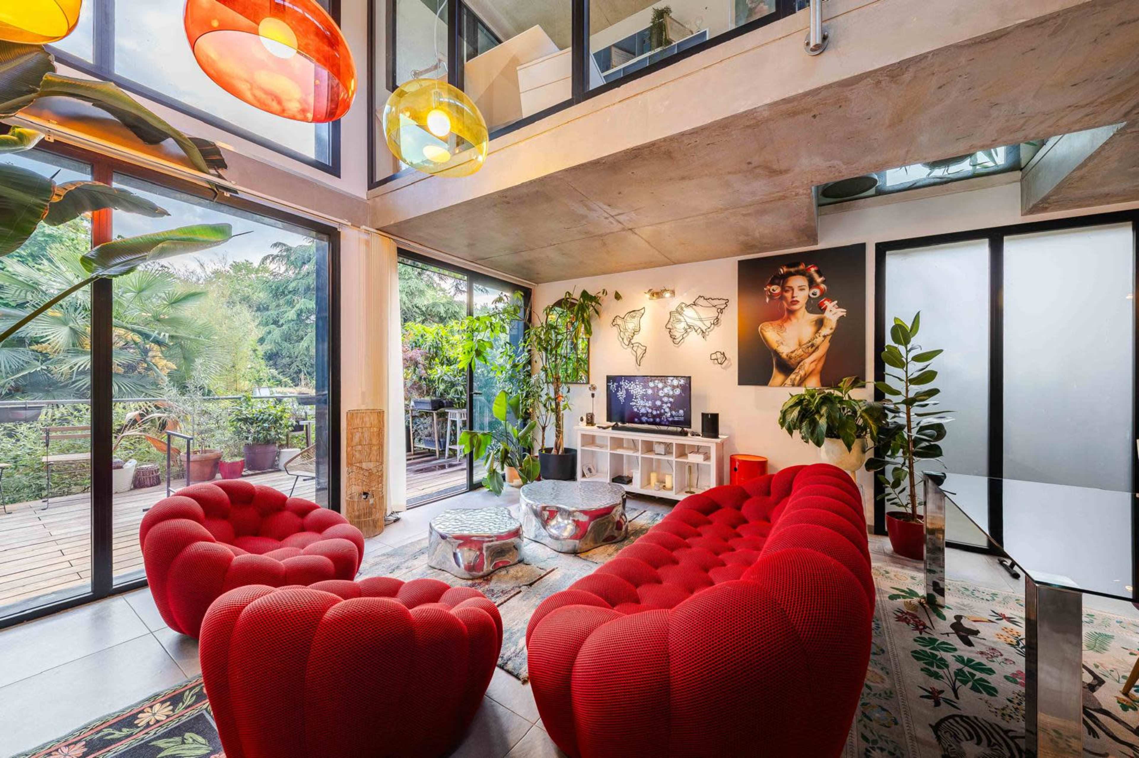 The image shows a modern living room featuring two red, plush armchairs, a glass coffee table, and large windows that open to a balcony surrounded by greenery.