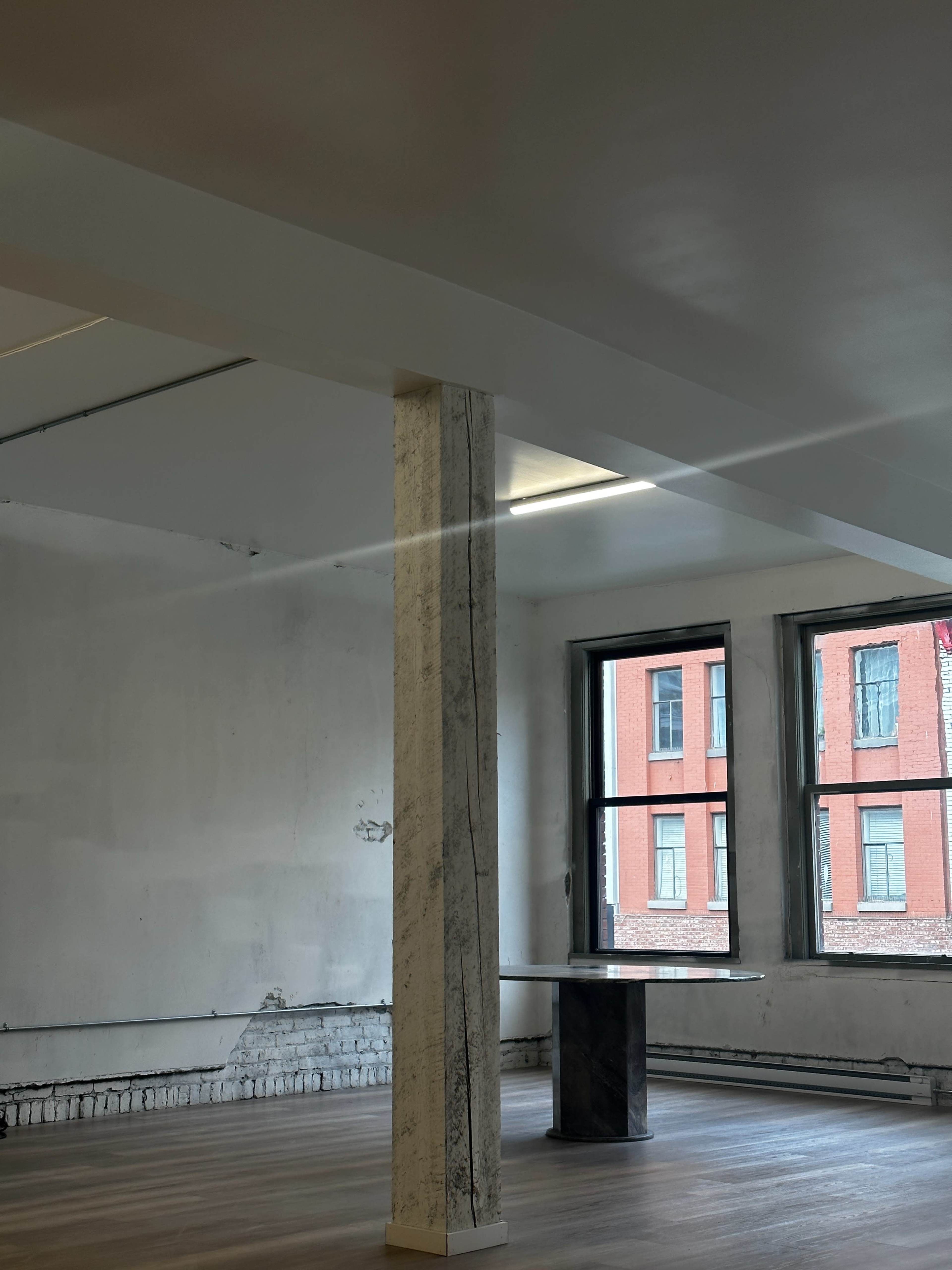 A spacious, partially empty room with a column and a table, featuring large windows that overlook a brick building.