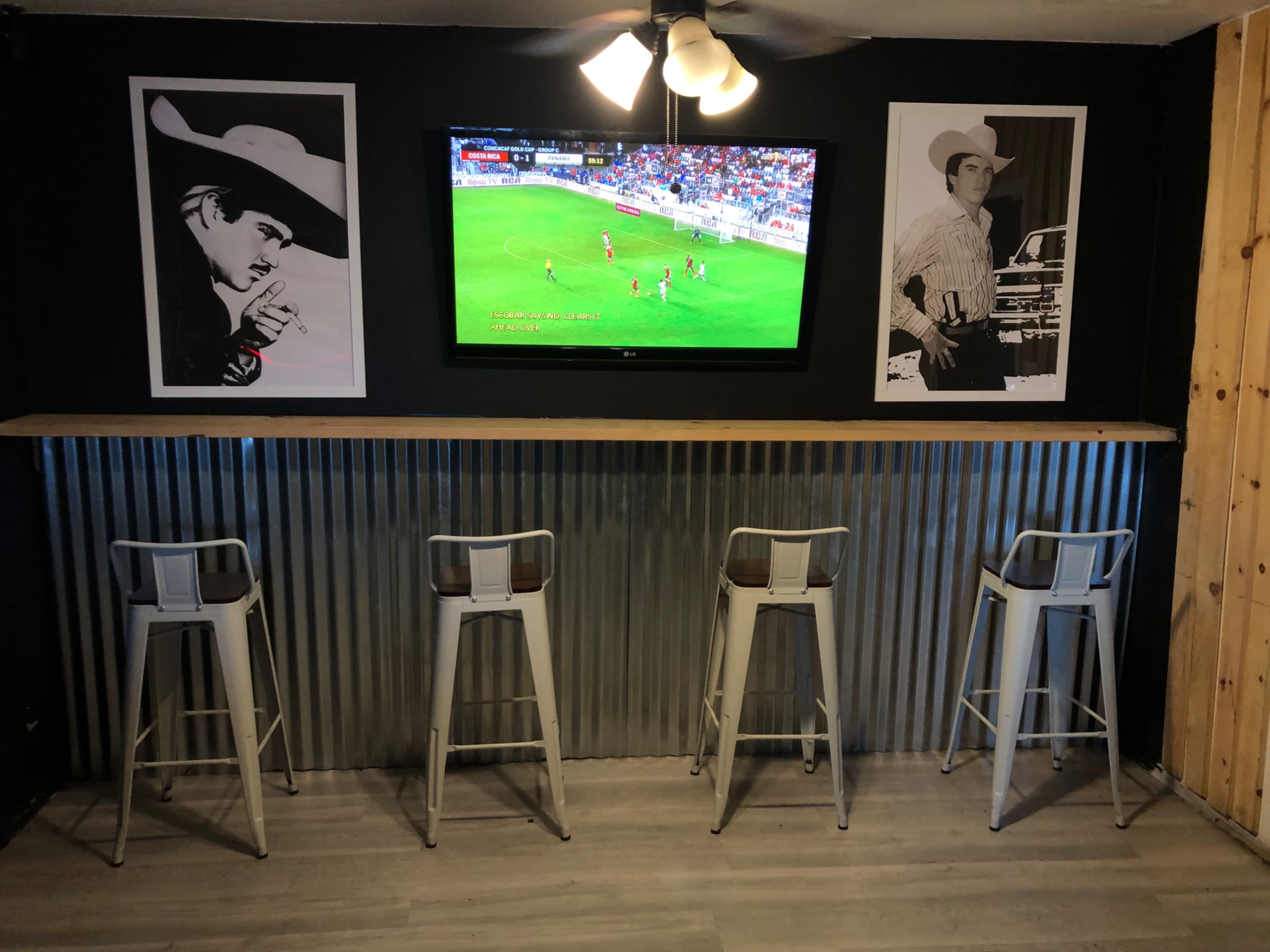 A bar area with four metal stools, a television displaying a soccer match, and two framed black-and-white photos on the wall.