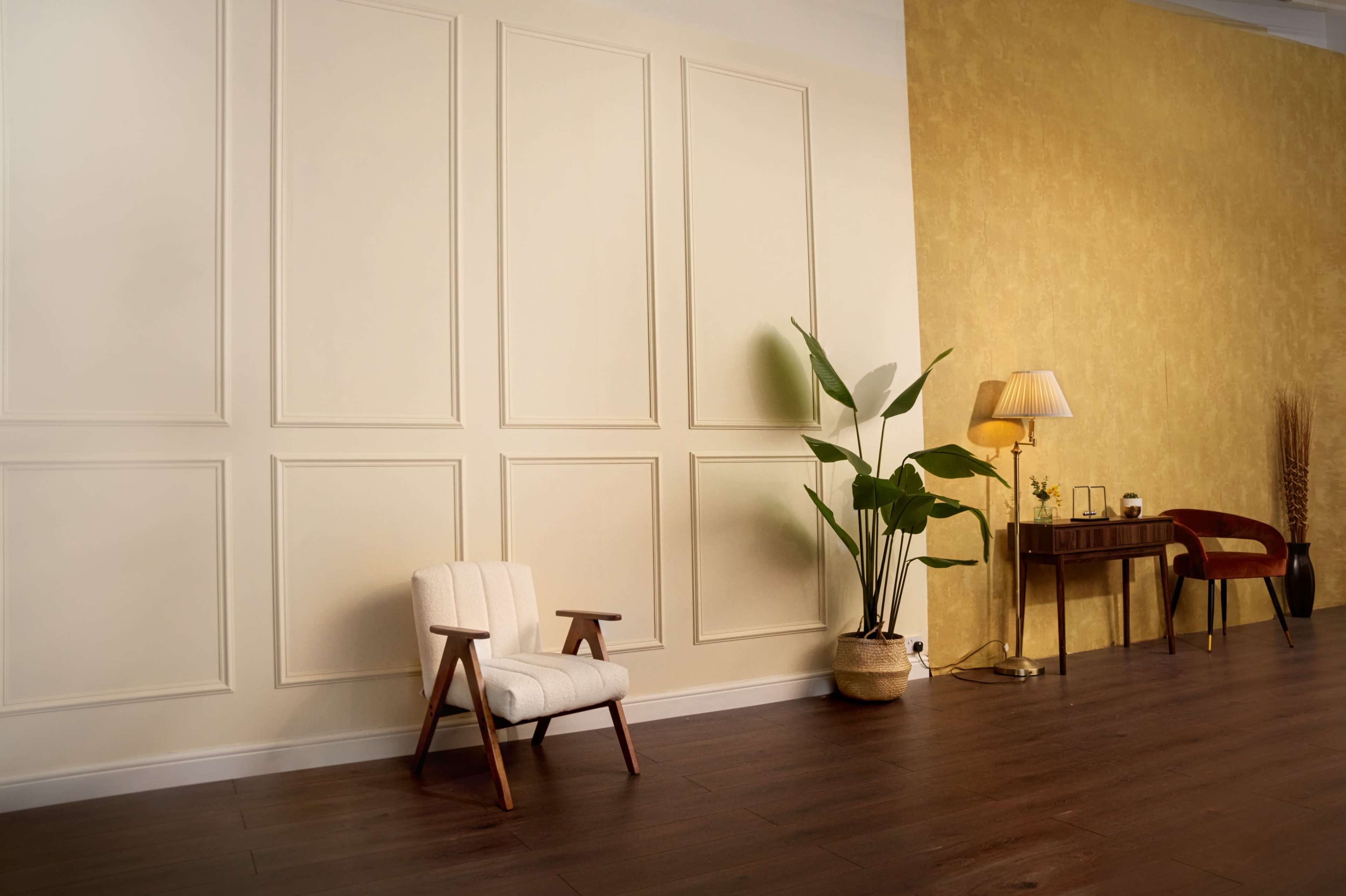 The image shows a minimalist interior space featuring a light-colored wall with paneling, a wooden chair, a potted plant, a small wooden table, and a lamp.
