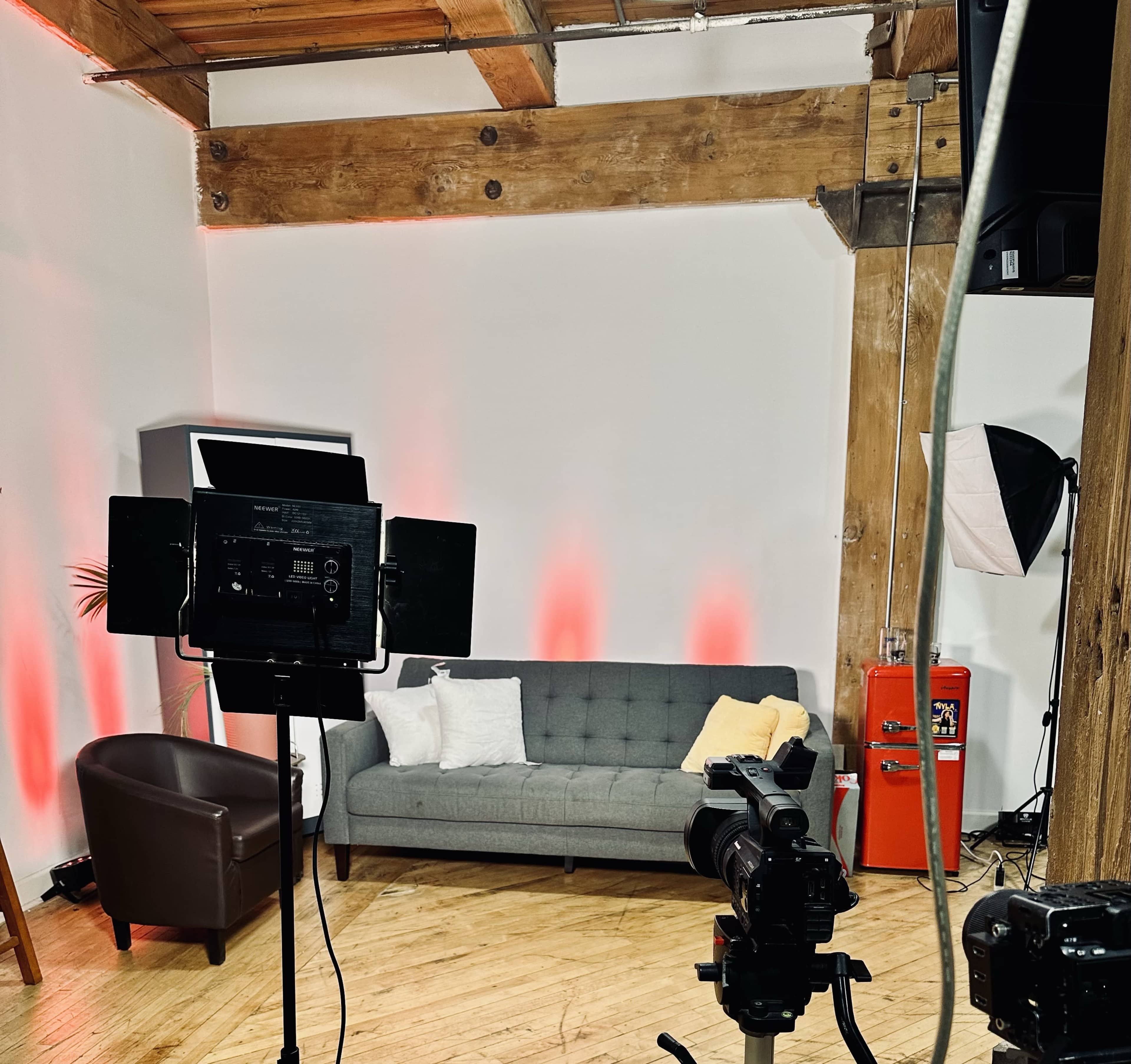 The image shows a cozy studio setup featuring a gray sofa, a brown armchair, various lighting equipment, and a vintage red fire extinguisher against a wooden backdrop.