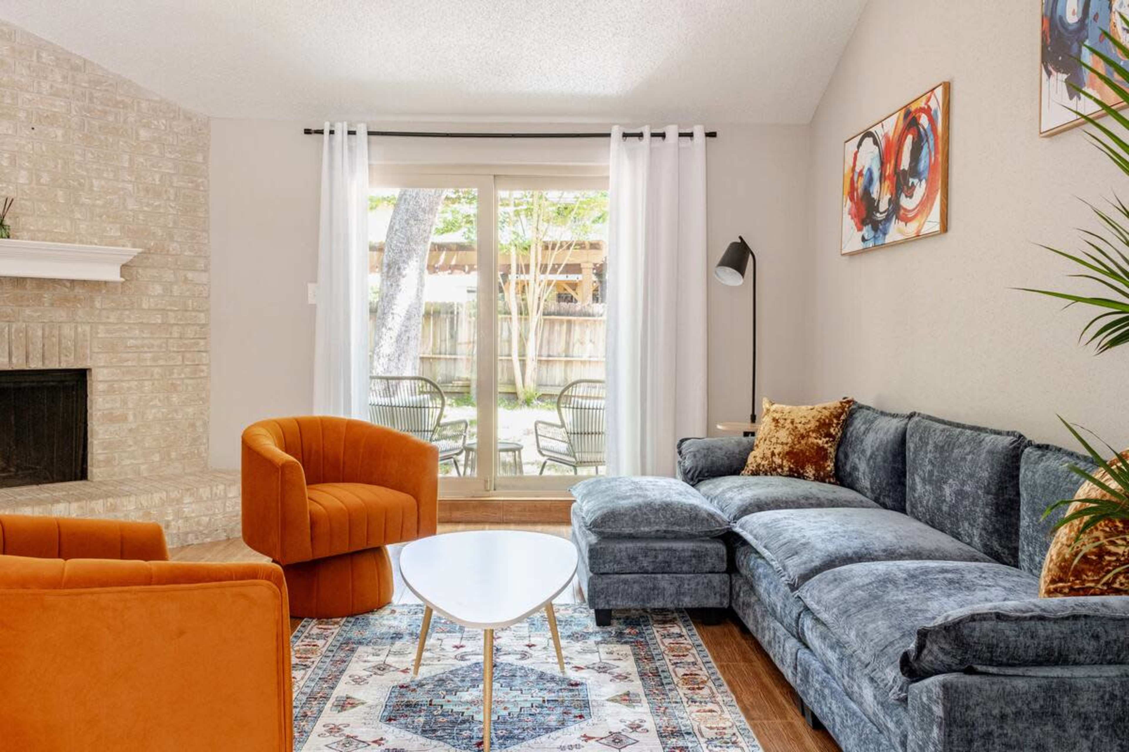 A modern living room features a blue sectional sofa, orange accent chairs, a round coffee table, and large windows overlooking a backyard.