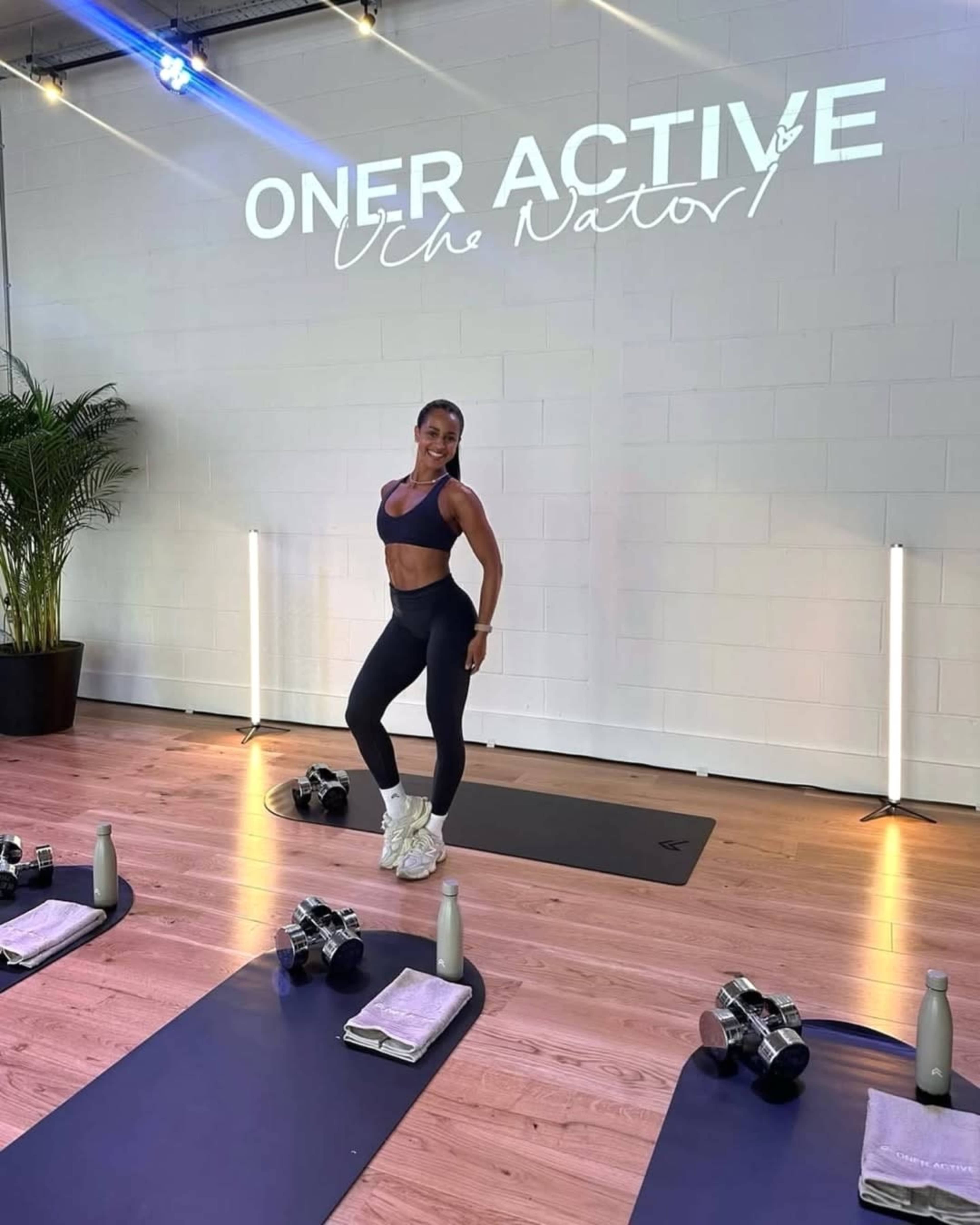 A fitness instructor poses in a brightly lit studio with exercise mats and weights arranged neatly on the floor.