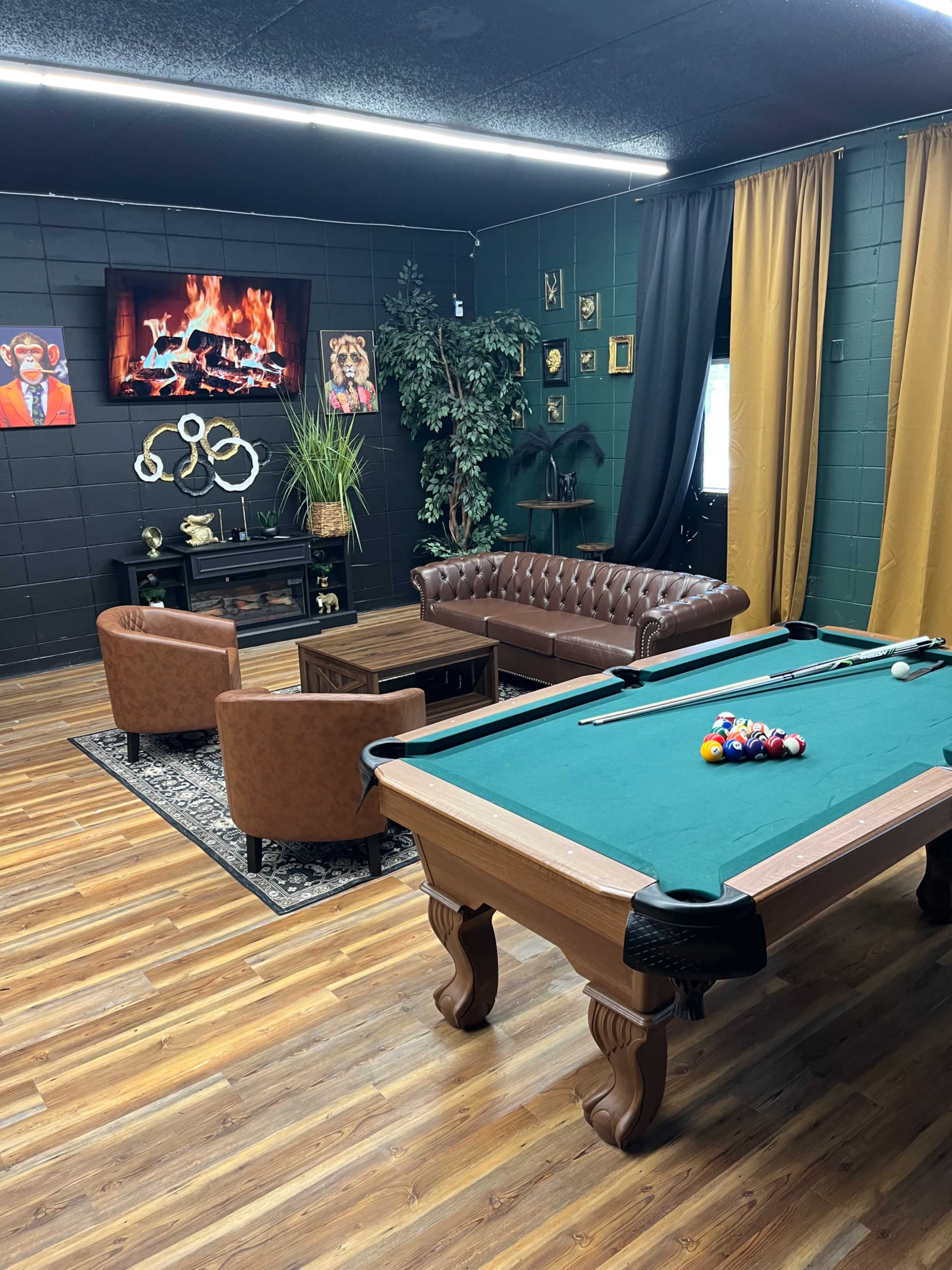 The image shows a cozy game room featuring a green felt pool table, a leather sofa and chairs, a fireplace on the wall, and various artworks displayed.