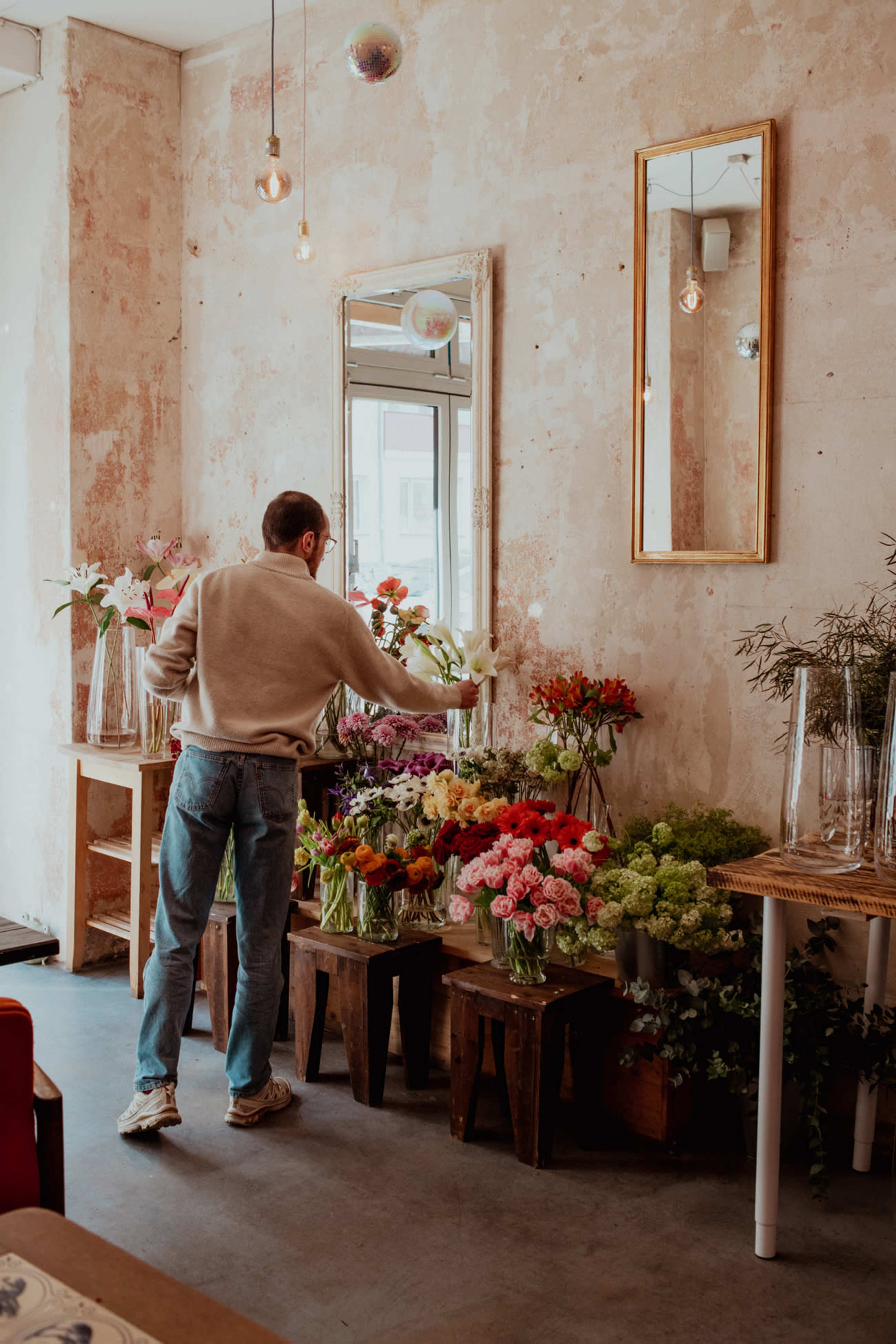 A person arranges flowers in a shop with a rustic interior featuring exposed walls and large mirrors.
