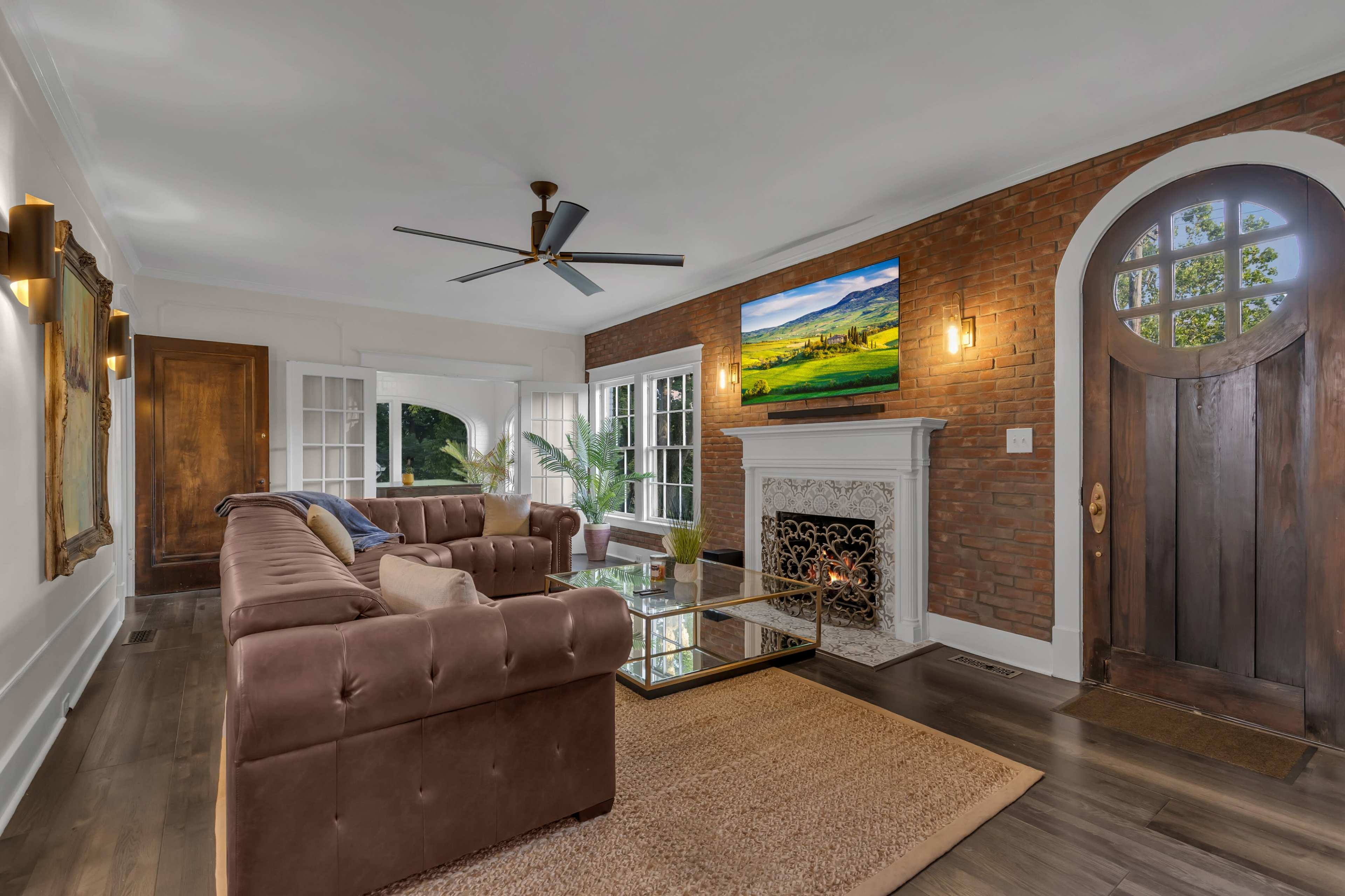 The living room features a brown sectional sofa, a glass coffee table, a TV mounted above a white fireplace, and a large wooden front door.