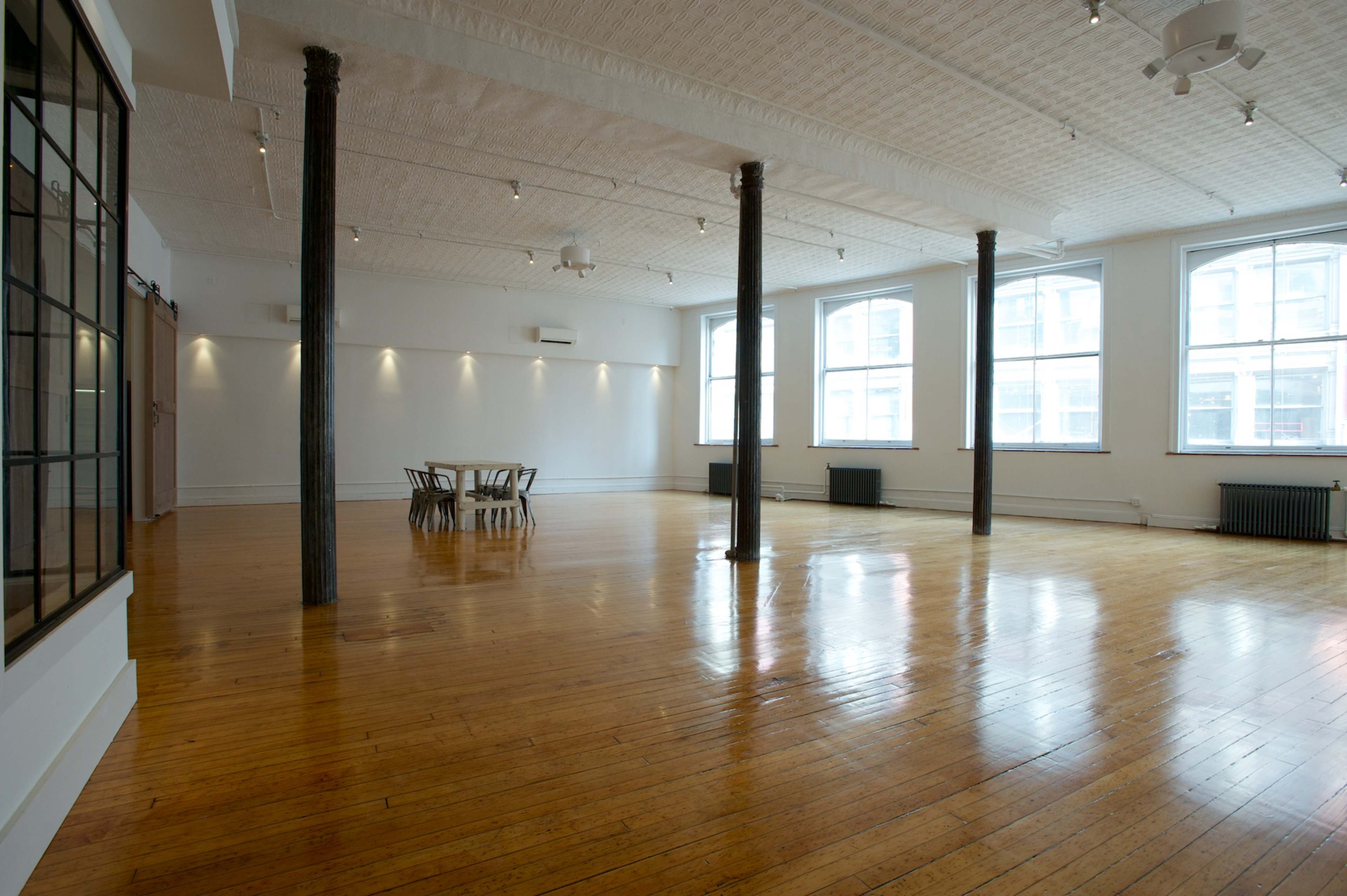 A spacious, empty room with wooden flooring, large windows, and exposed structural columns.