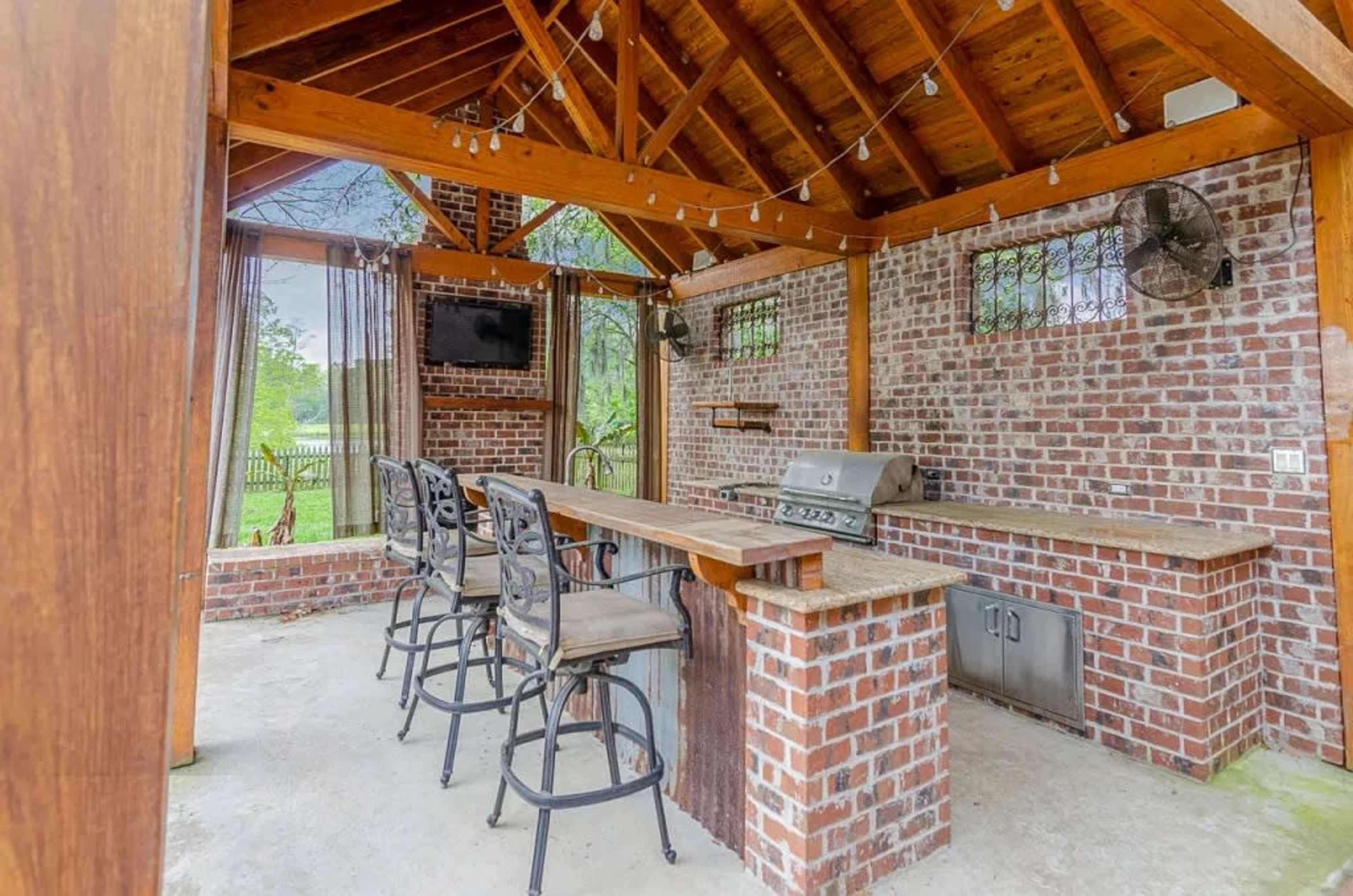The image shows an outdoor kitchen space with a brick structure, a grilling area, high chairs, and hanging lights.