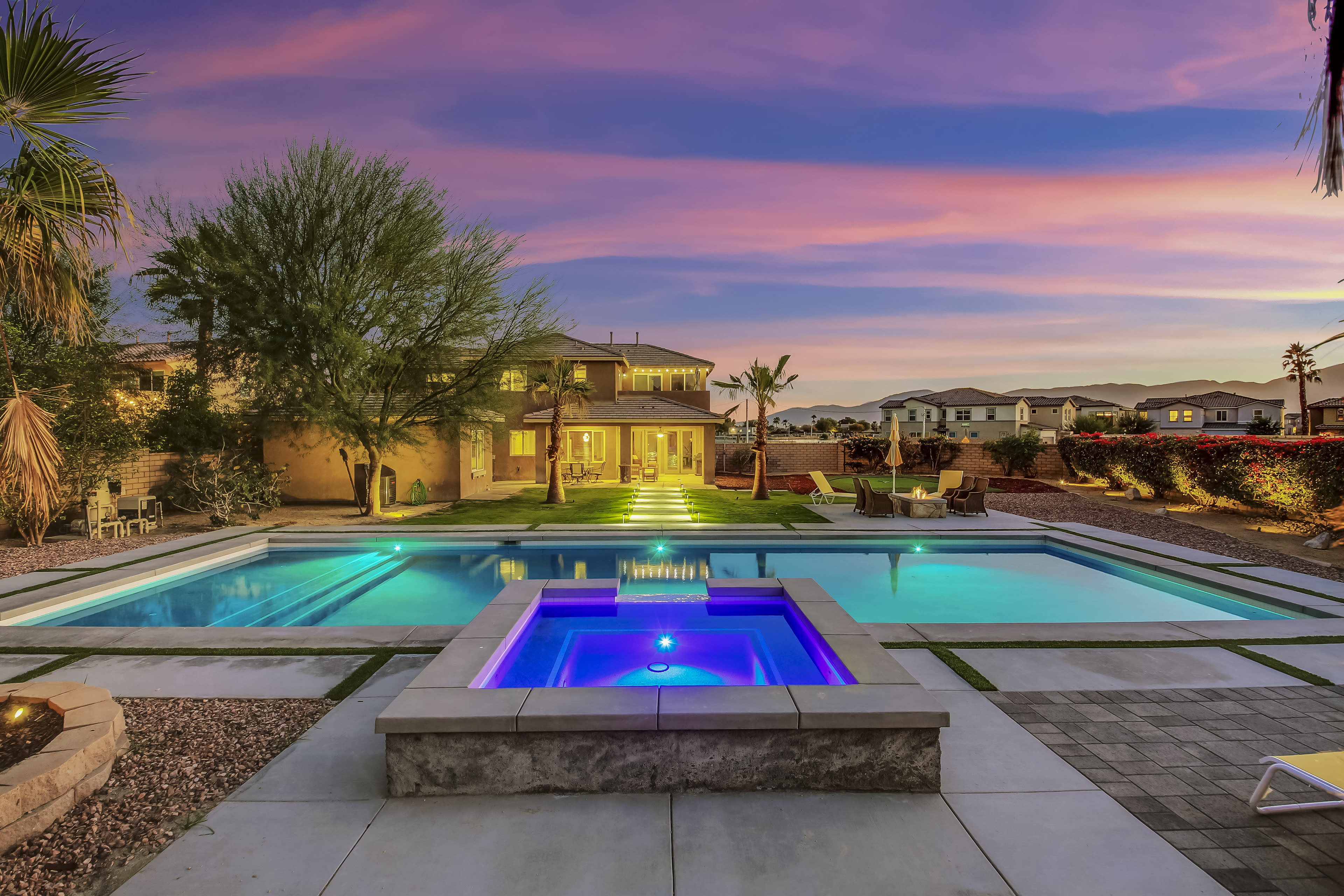 A backyard features a large, illuminated swimming pool with a raised spa, surrounded by landscaping and a two-story house in the background under a colorful sunset.