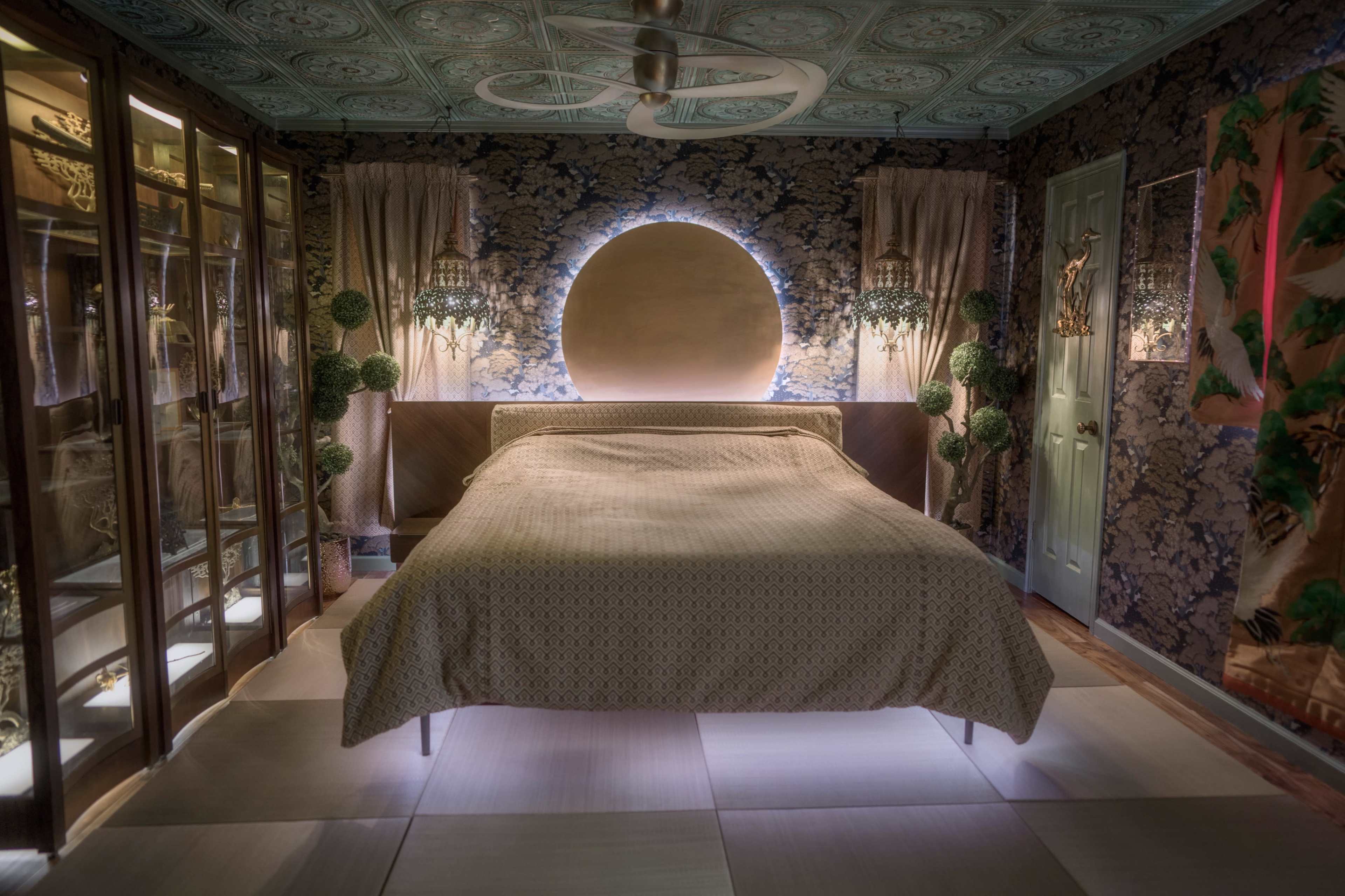 The image shows a dimly lit bedroom with a large circular feature above the bed, intricate wall designs, and glass cabinets flanking the space.