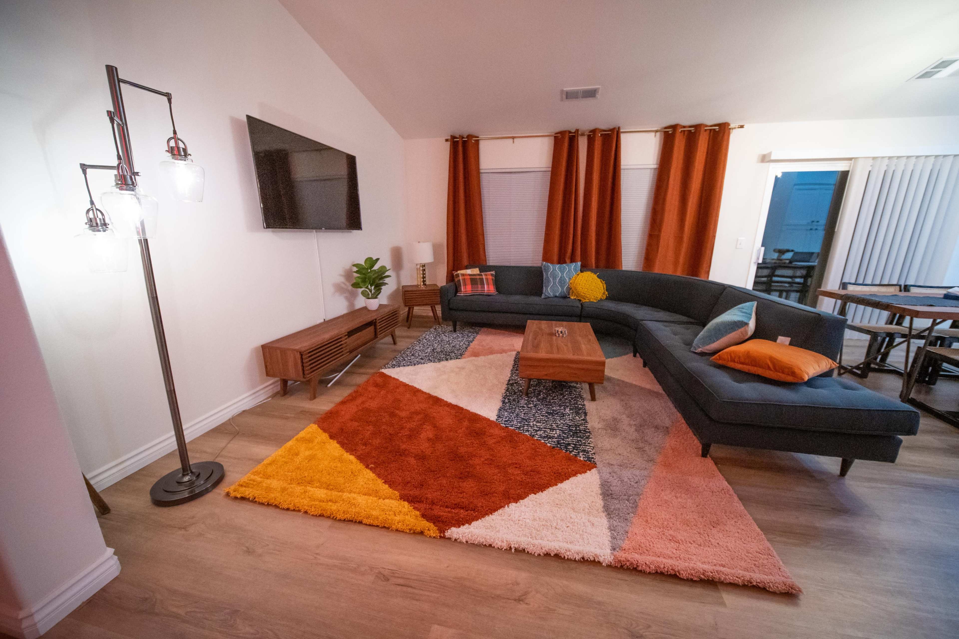 The living room features a large sectional sofa, a coffee table, and a colorful geometric area rug under a bright light fixture.