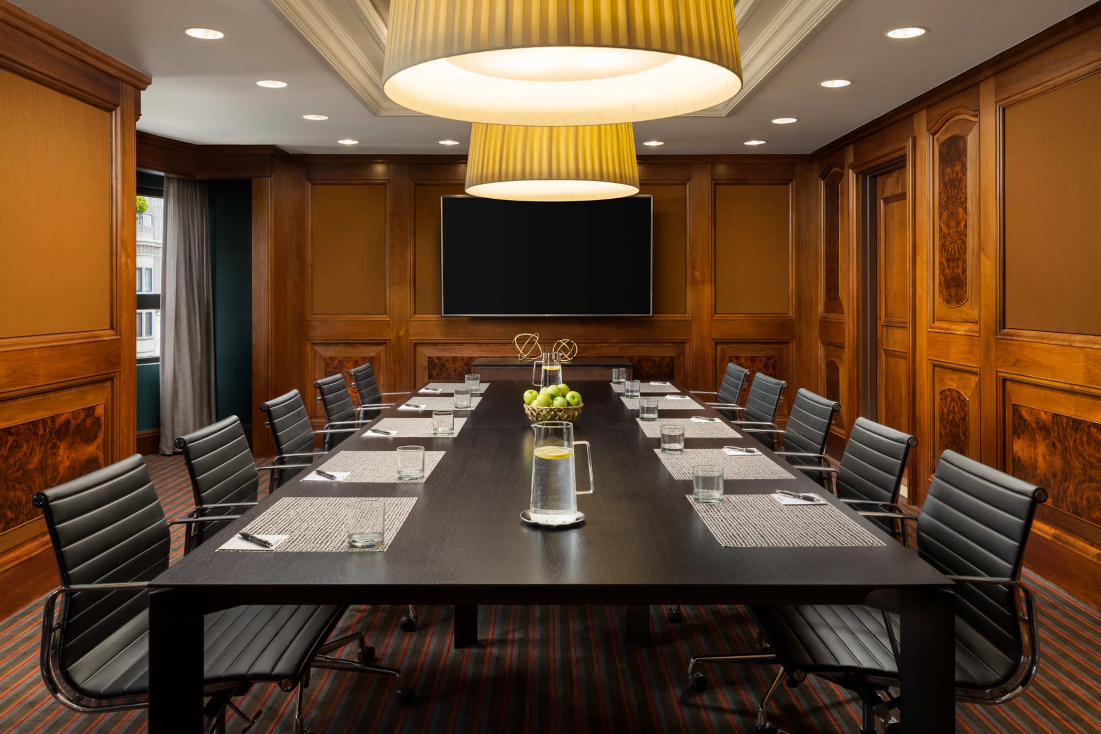 A conference room with a large wooden table surrounded by black chairs, featuring a modern lighting fixture and a large screen on the wall.
