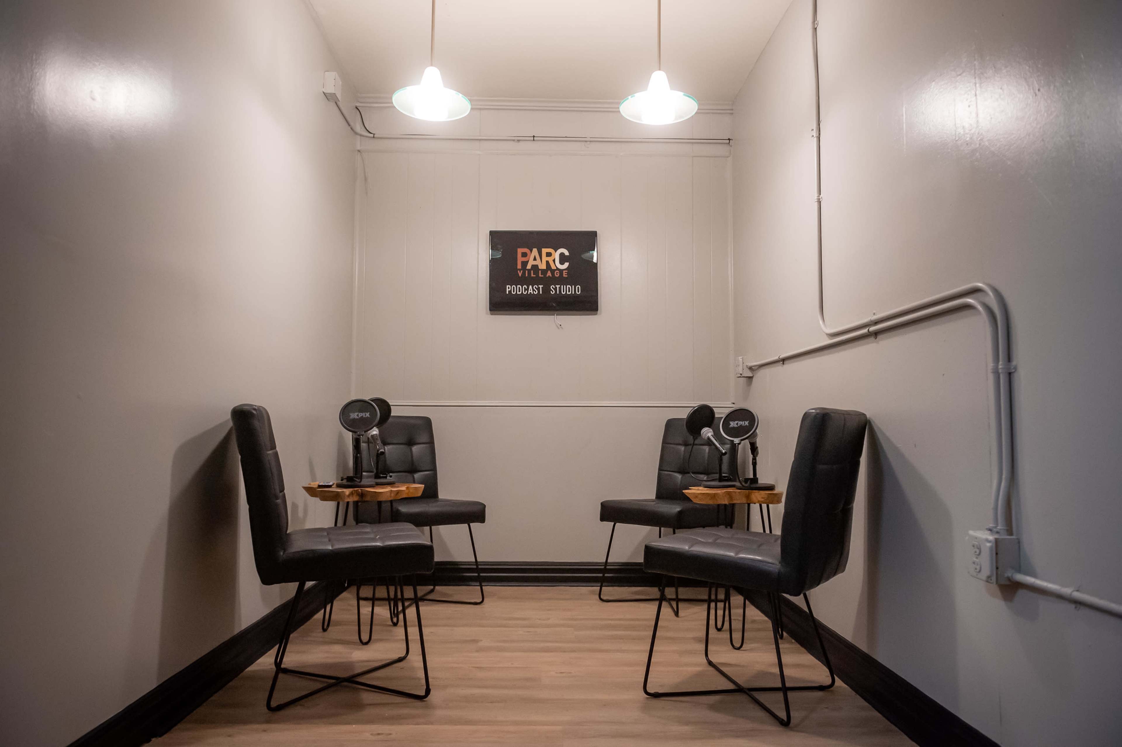 The image shows a minimalist podcast studio featuring four black chairs arranged around wooden side tables under soft ceiling lights.