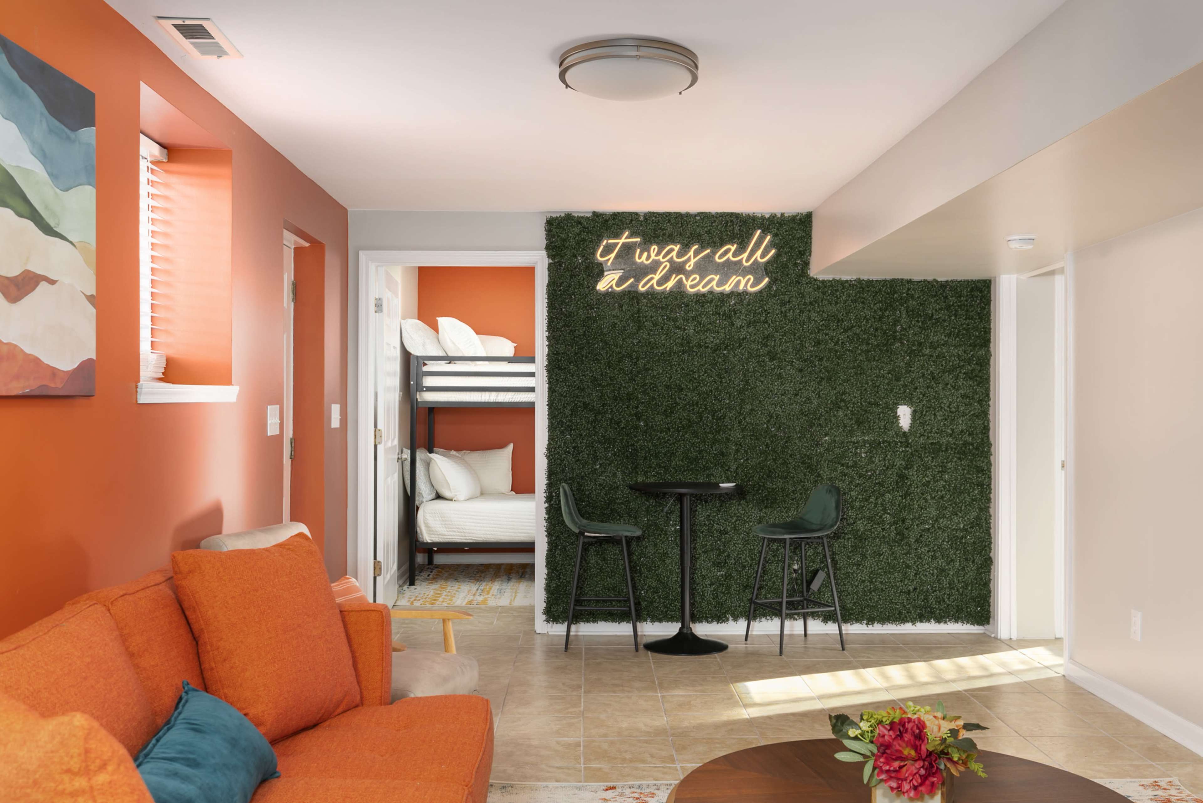 A well-lit modern living space features an orange sofa, a small dining table, and a green wall adorned with the phrase "I was all a dream."