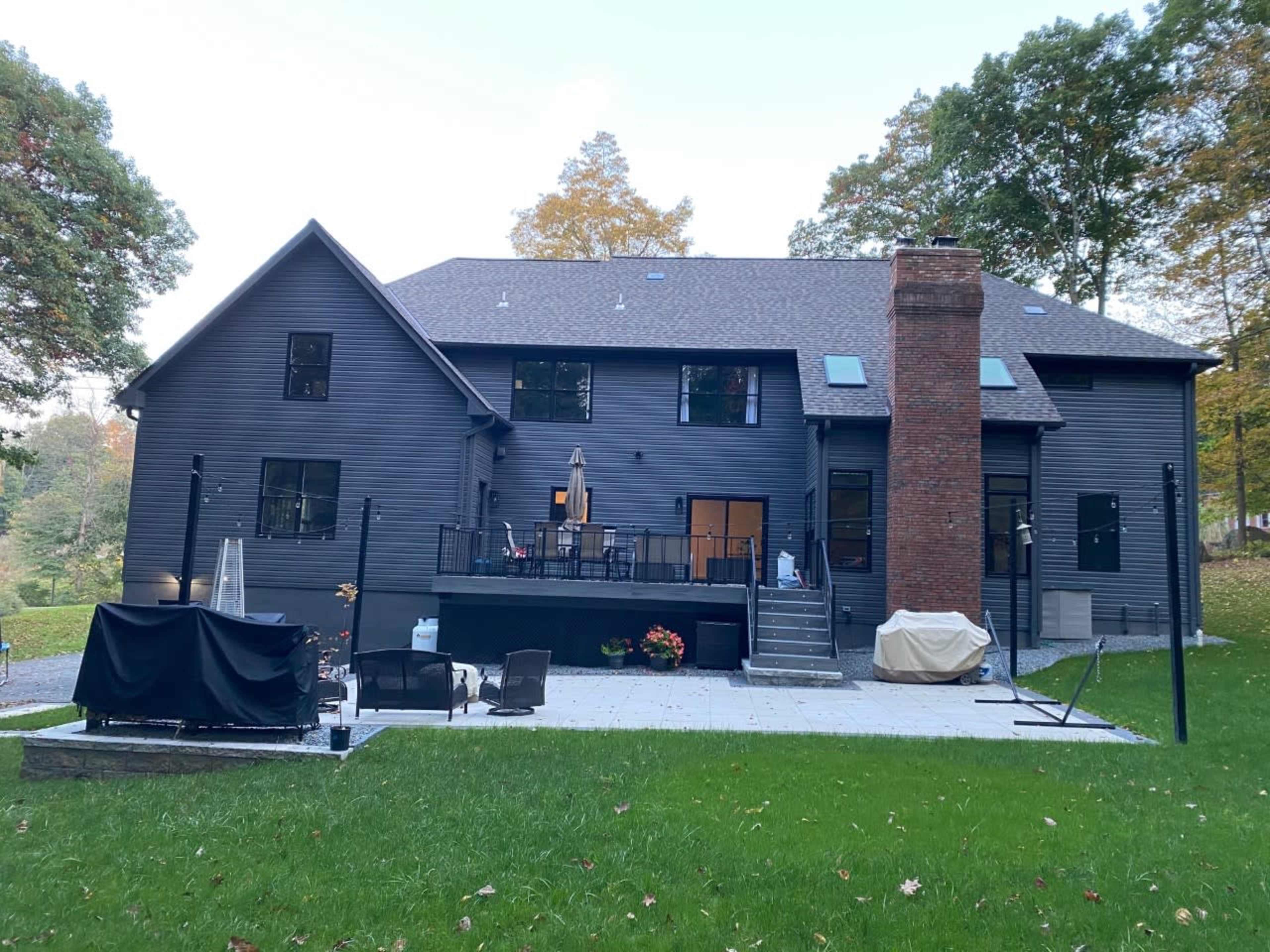A modern two-story house with a dark exterior features a large patio, outdoor seating area, and a chimney.