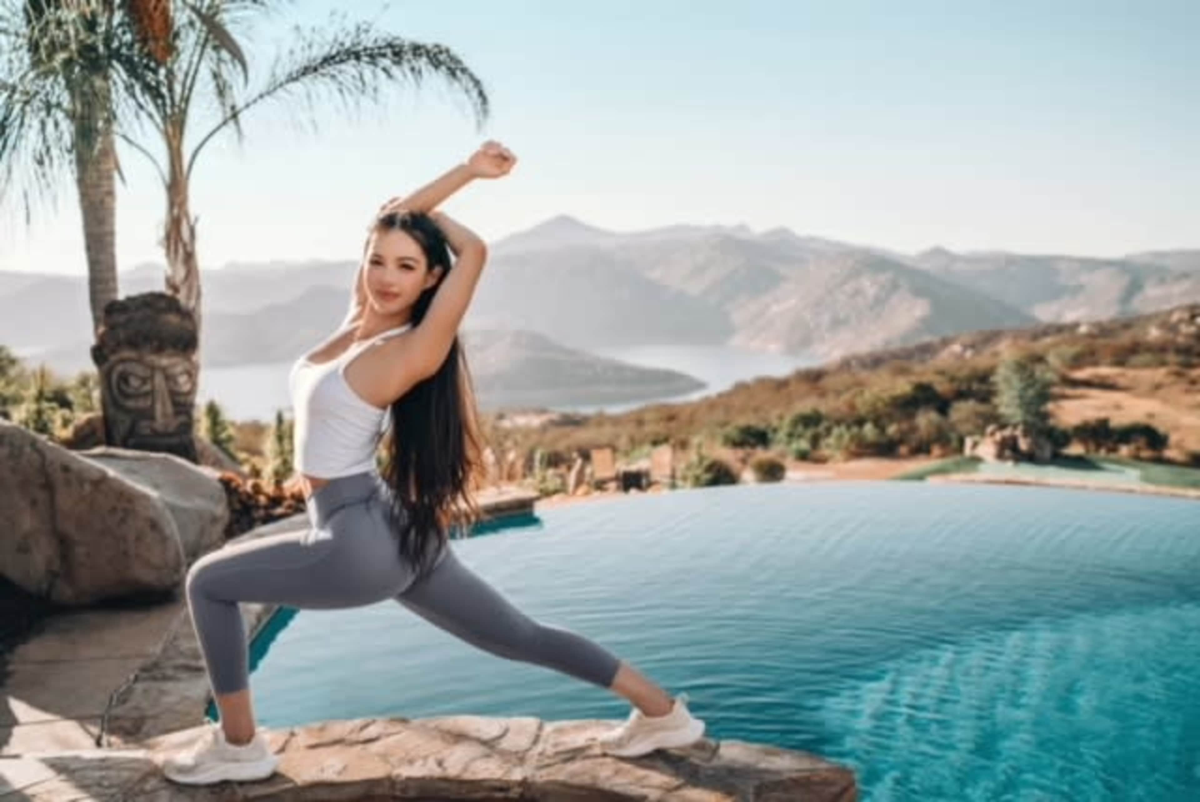 A woman in athletic wear stretches by a pool overlooking a mountainous landscape.