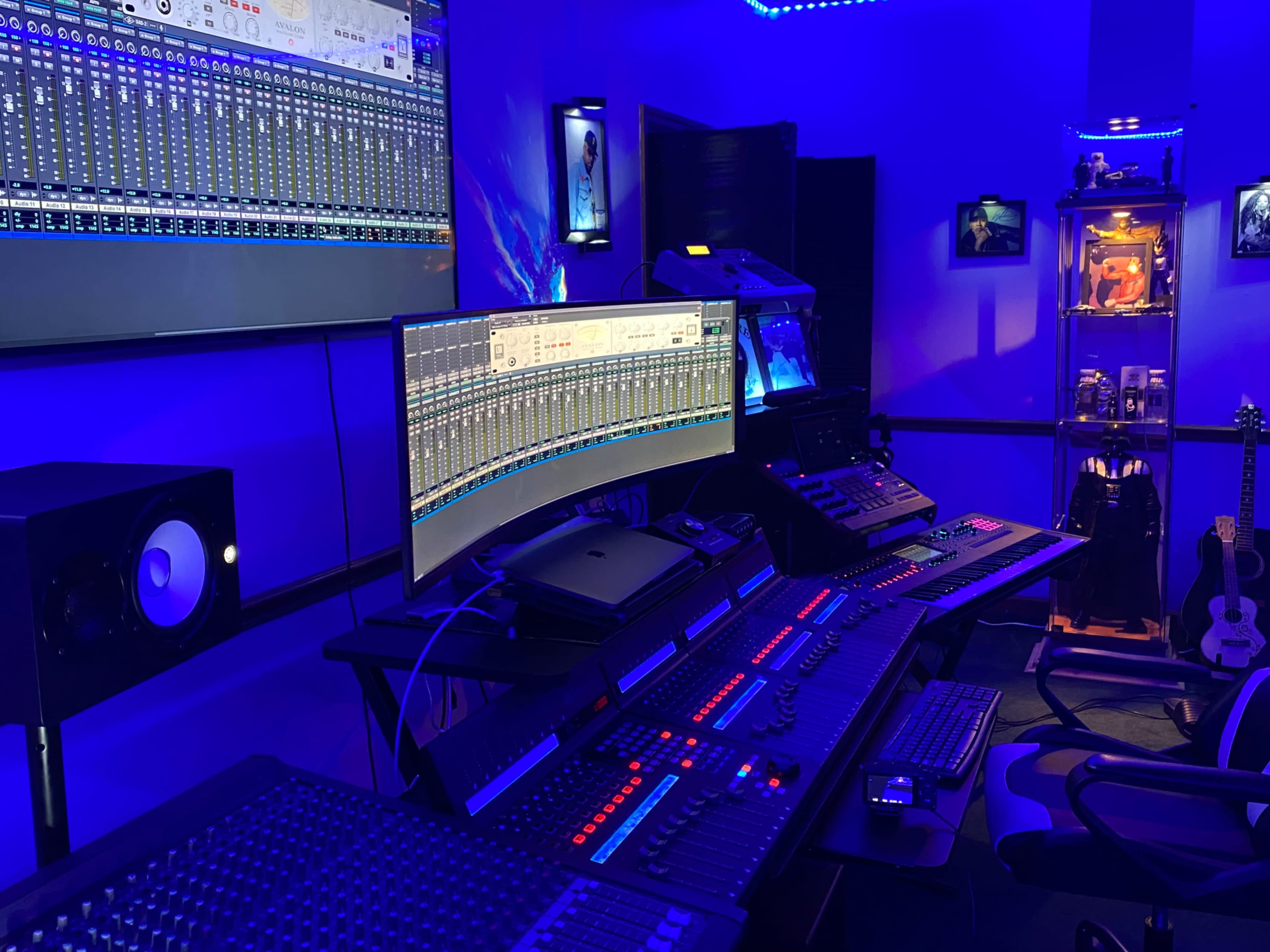 A modern music studio with multiple screens displaying audio tracks, a central mixing console with illuminated controls, and blue LED lighting throughout the room.