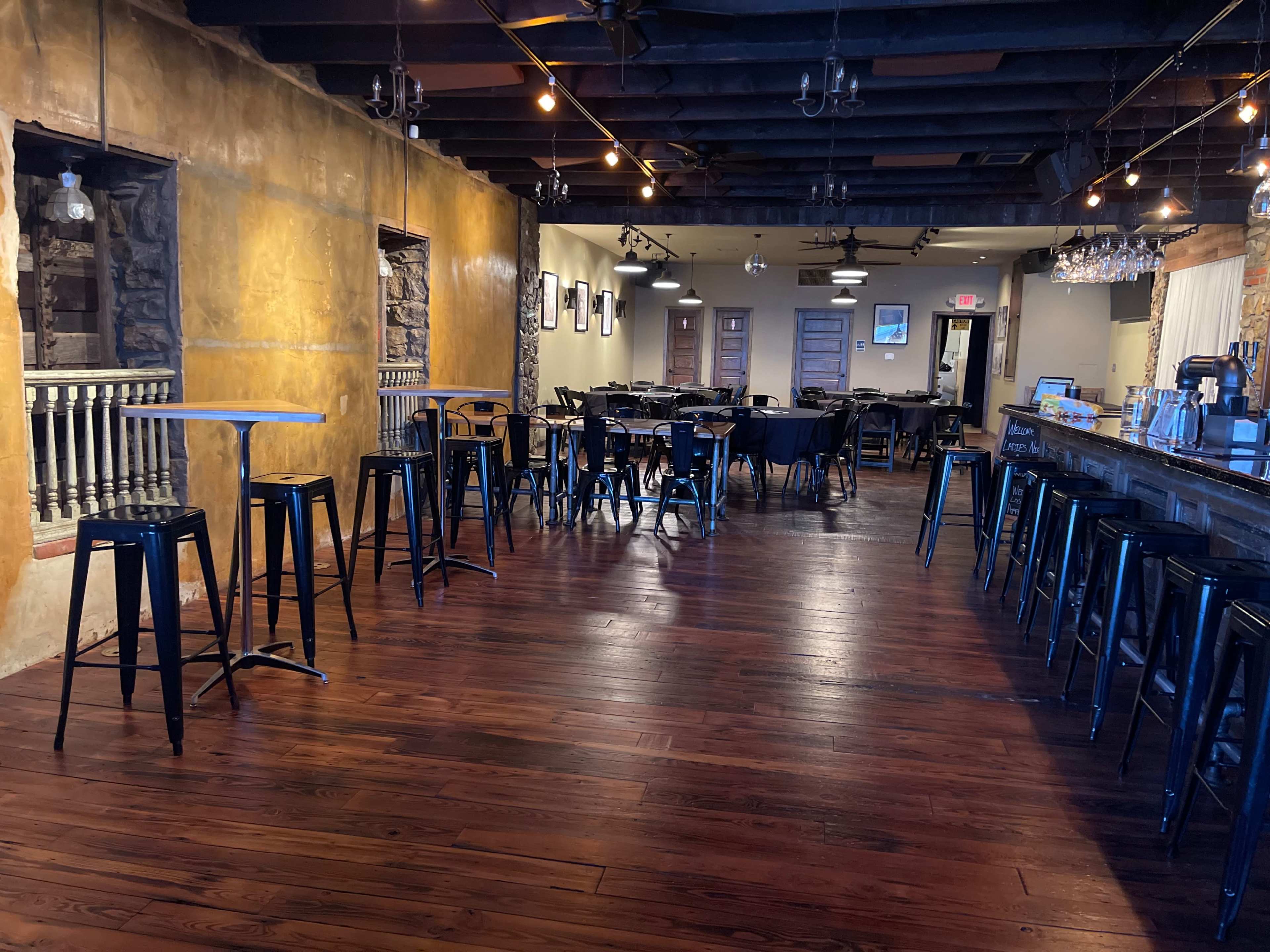A spacious restaurant interior featuring dark wooden floors, a bar with high stools, and tables set up for dining.