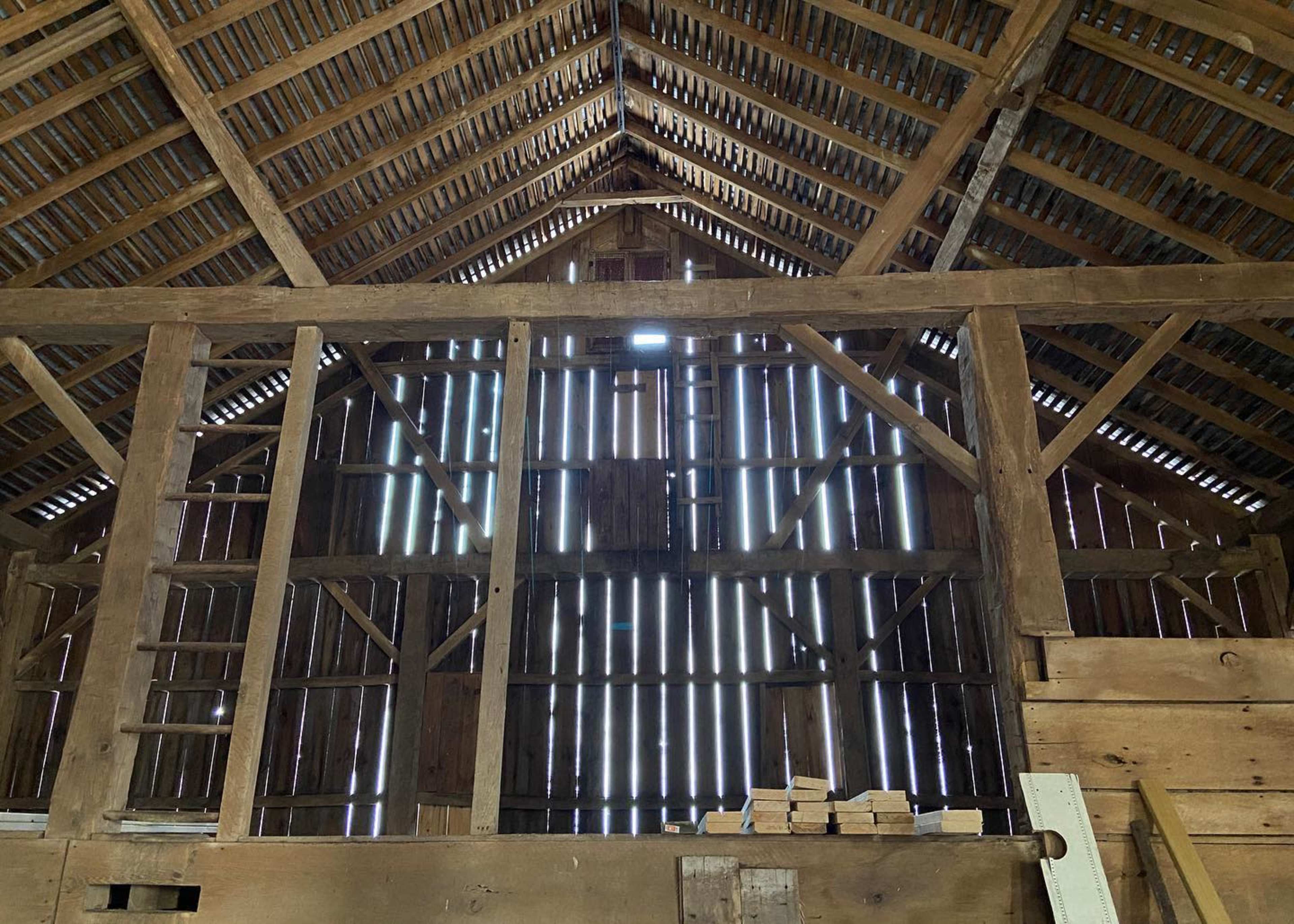 The interior of a wooden barn features a framework of beams and spaced slats allowing light to filter through.