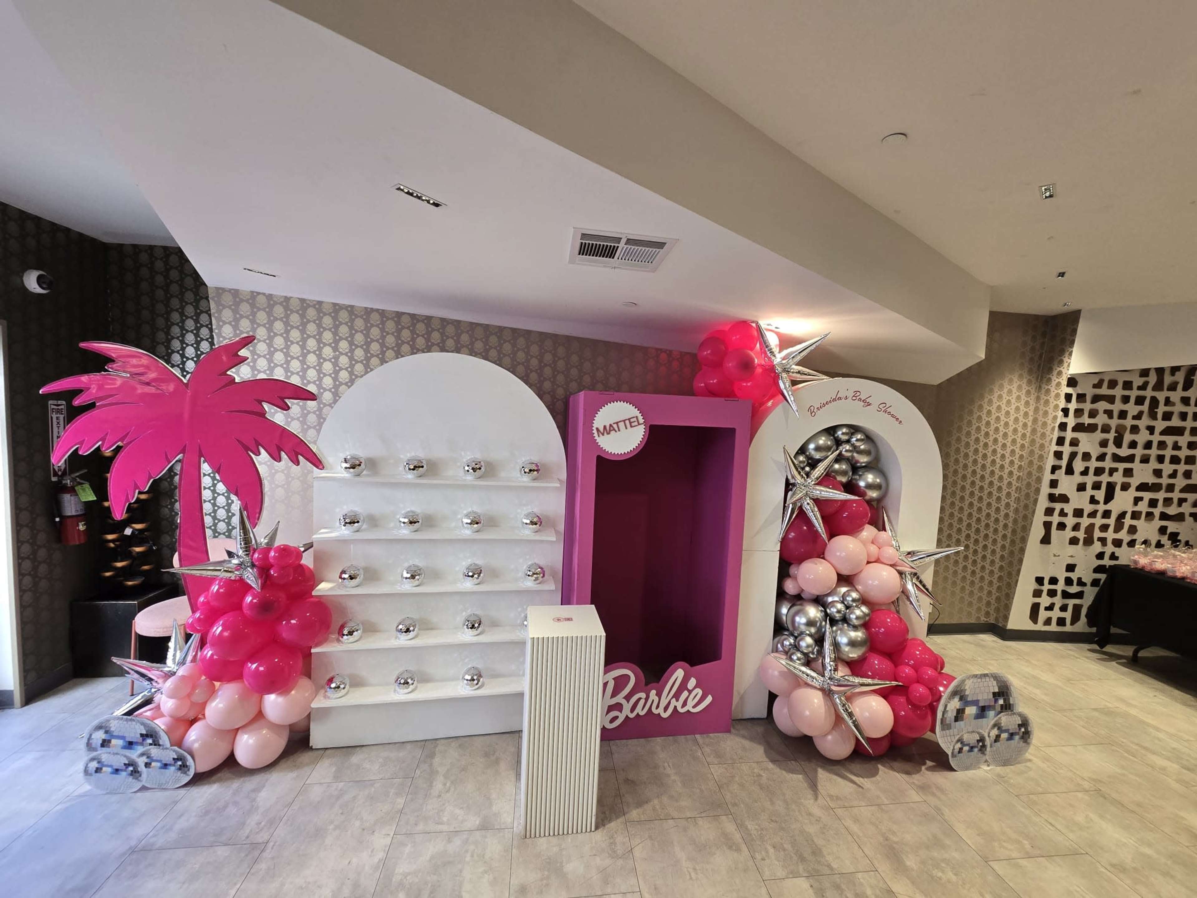 The scene features a brightly decorated area with a pink backdrop, white arches, and a combination of balloons in pink and silver, alongside a palm tree decoration.
