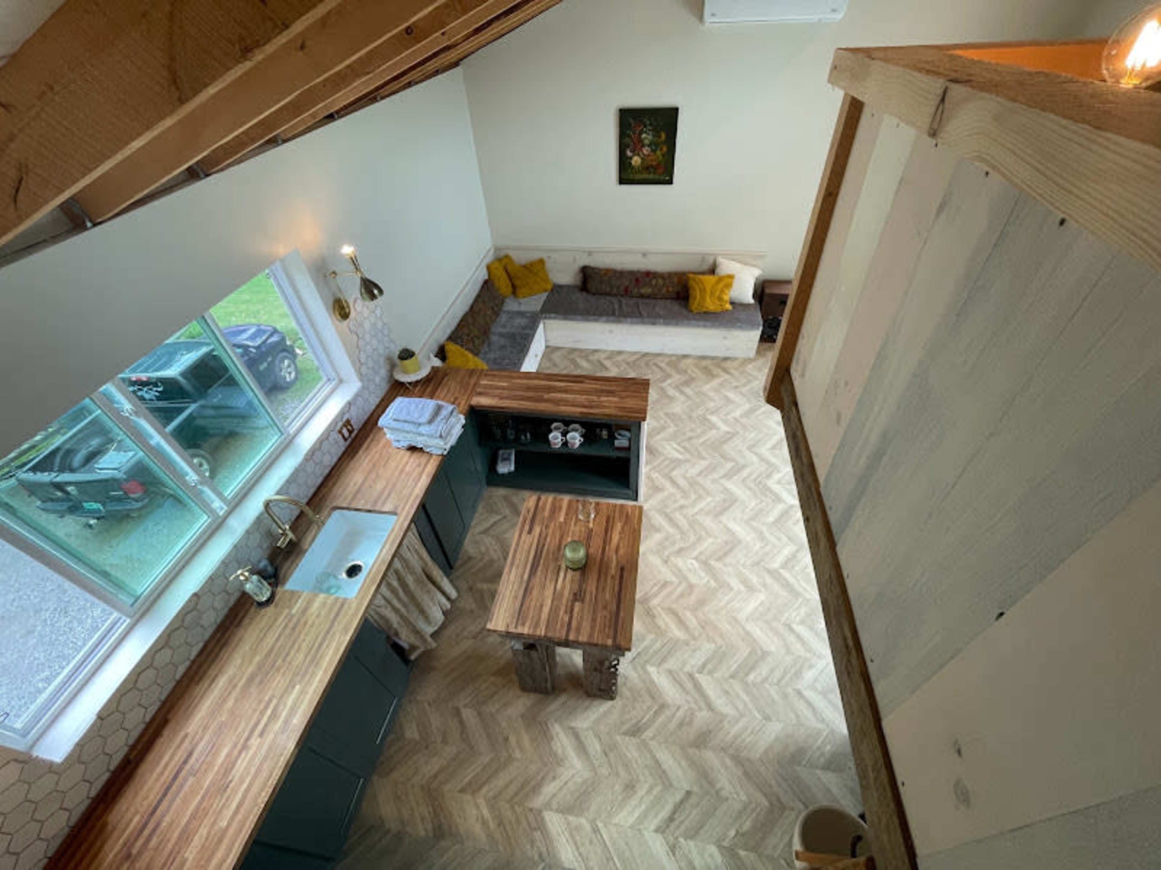 The image shows an open-concept kitchen and living area with wooden countertops, a herringbone-patterned floor, and a large window allowing natural light to enter.