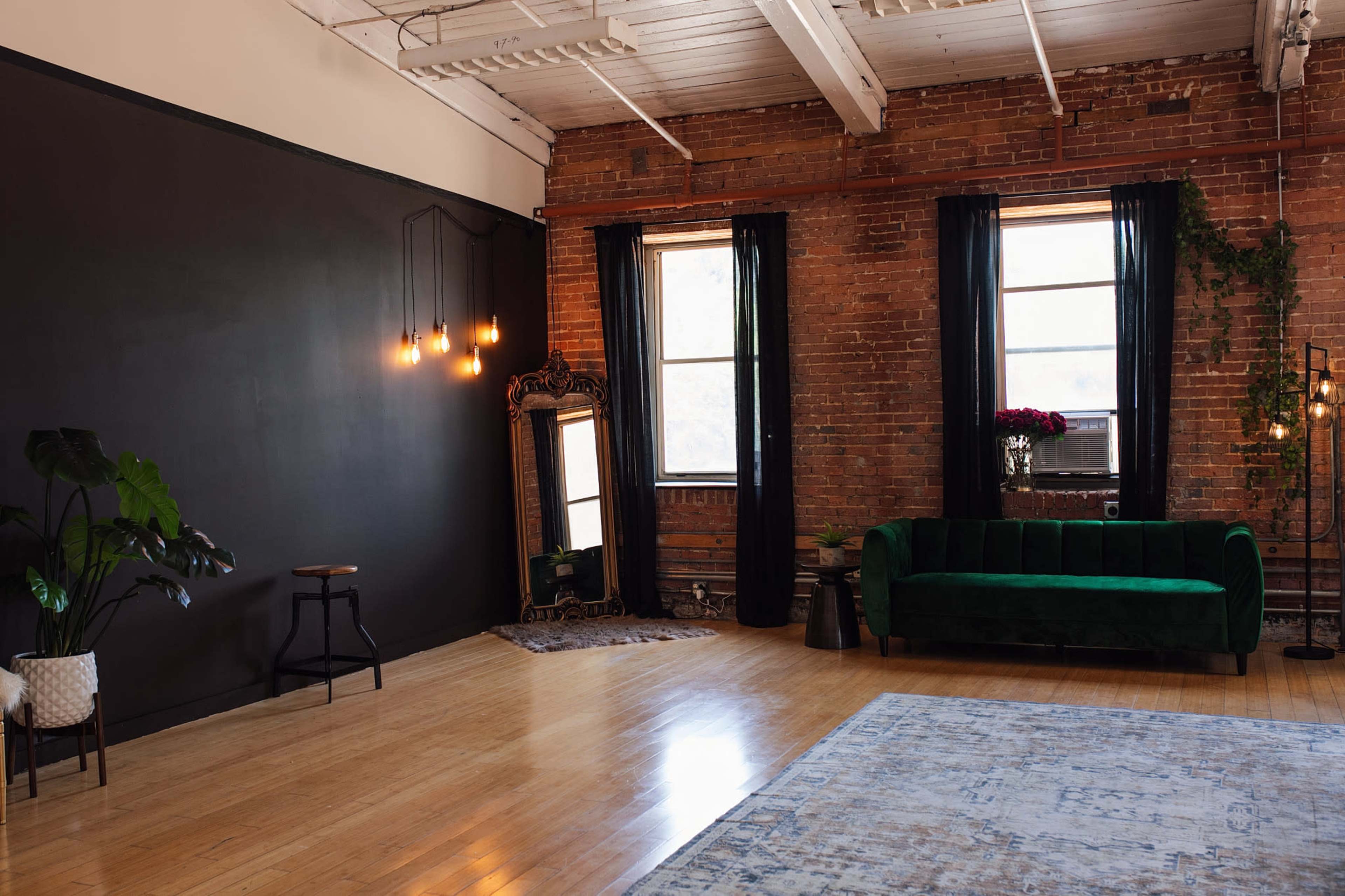 The image depicts a spacious room with exposed brick walls, large windows, a green velvet sofa, and a decorative mirror, complemented by hanging light bulbs and indoor plants.