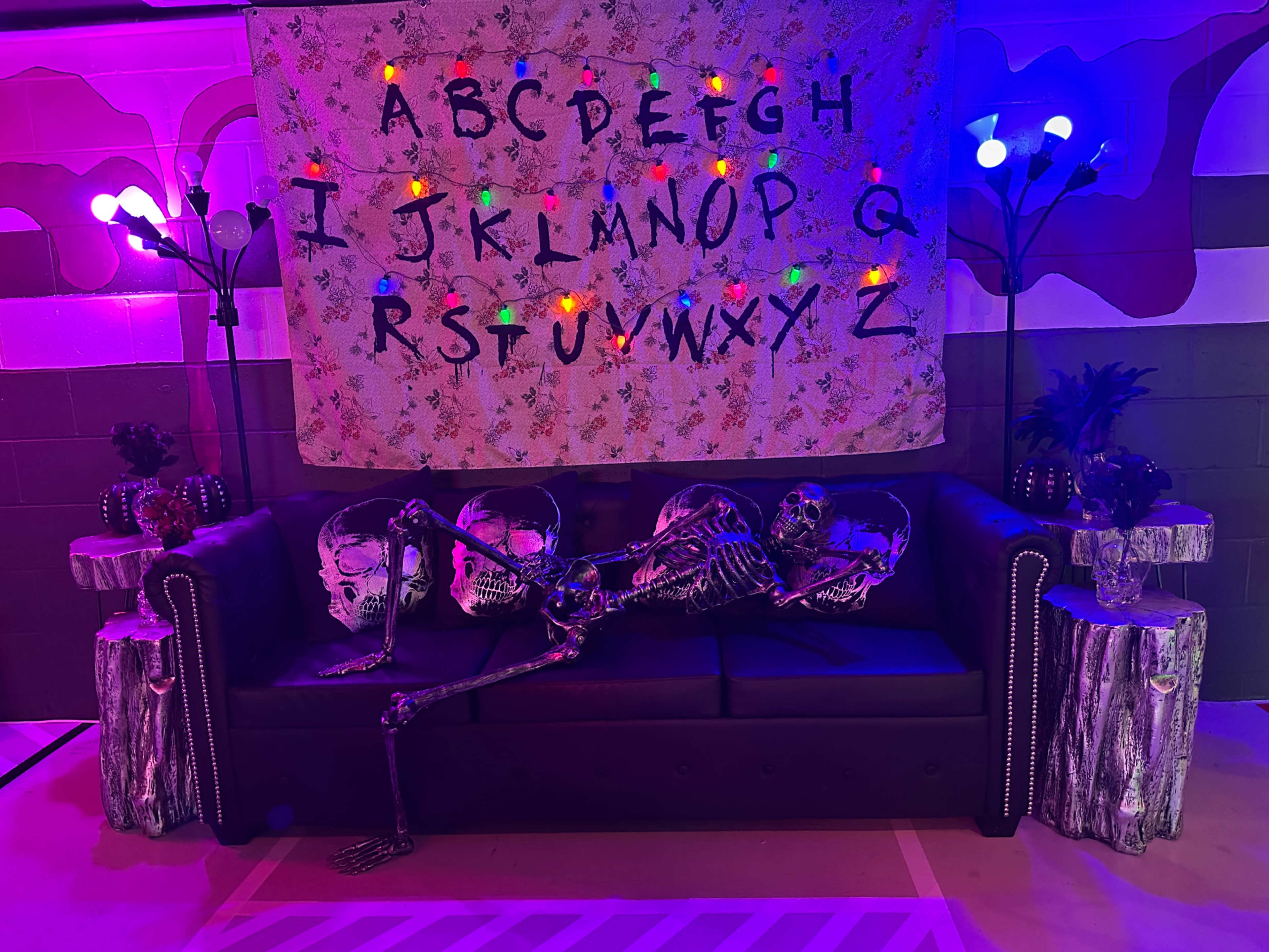 A skeleton is lounging on a black couch beneath a decorative banner of the alphabet, surrounded by colorful lights and pumpkins.