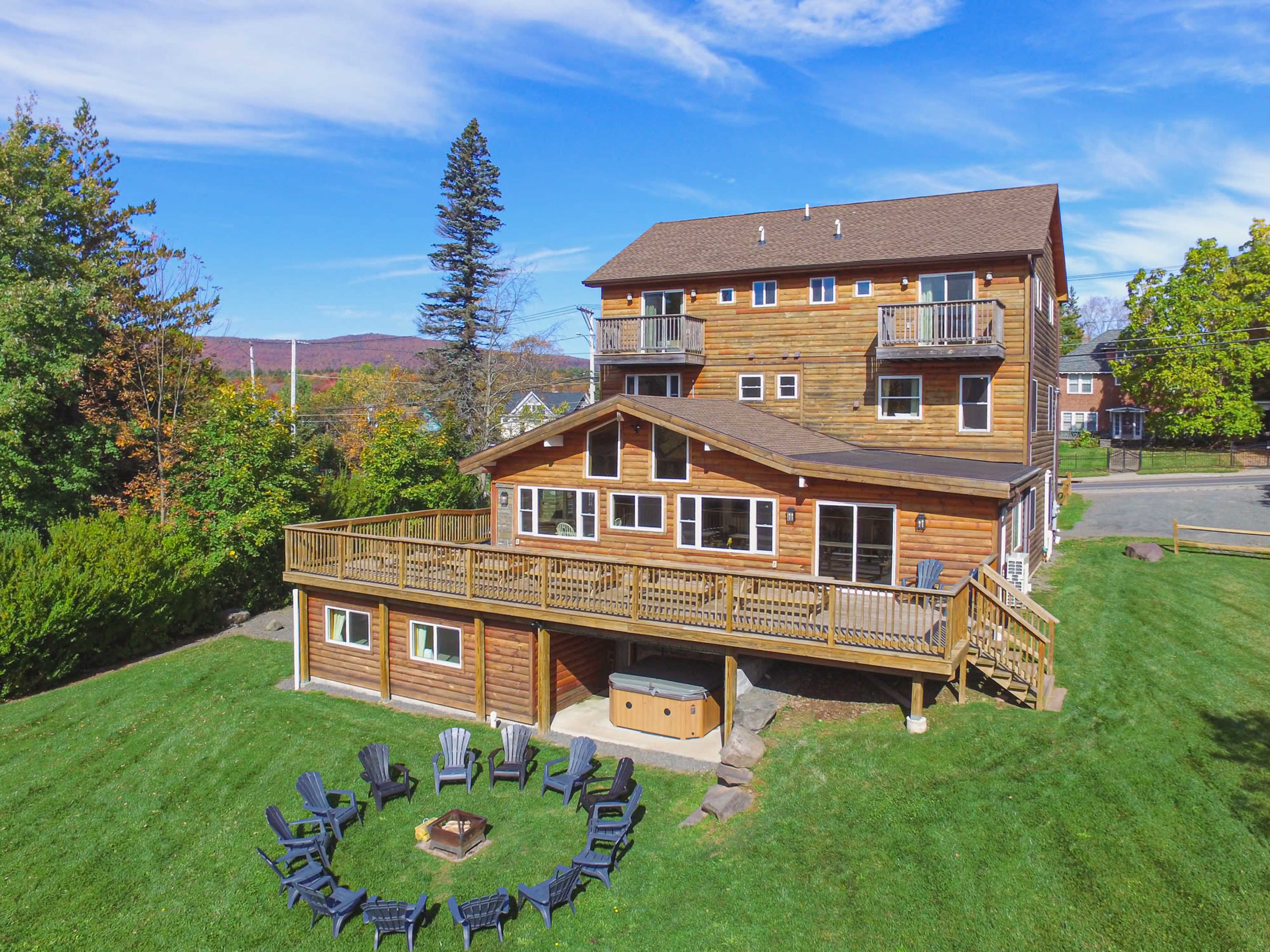 A multi-level wooden house with a spacious deck and a circular arrangement of outdoor seating surrounds a fire pit in a green yard.
