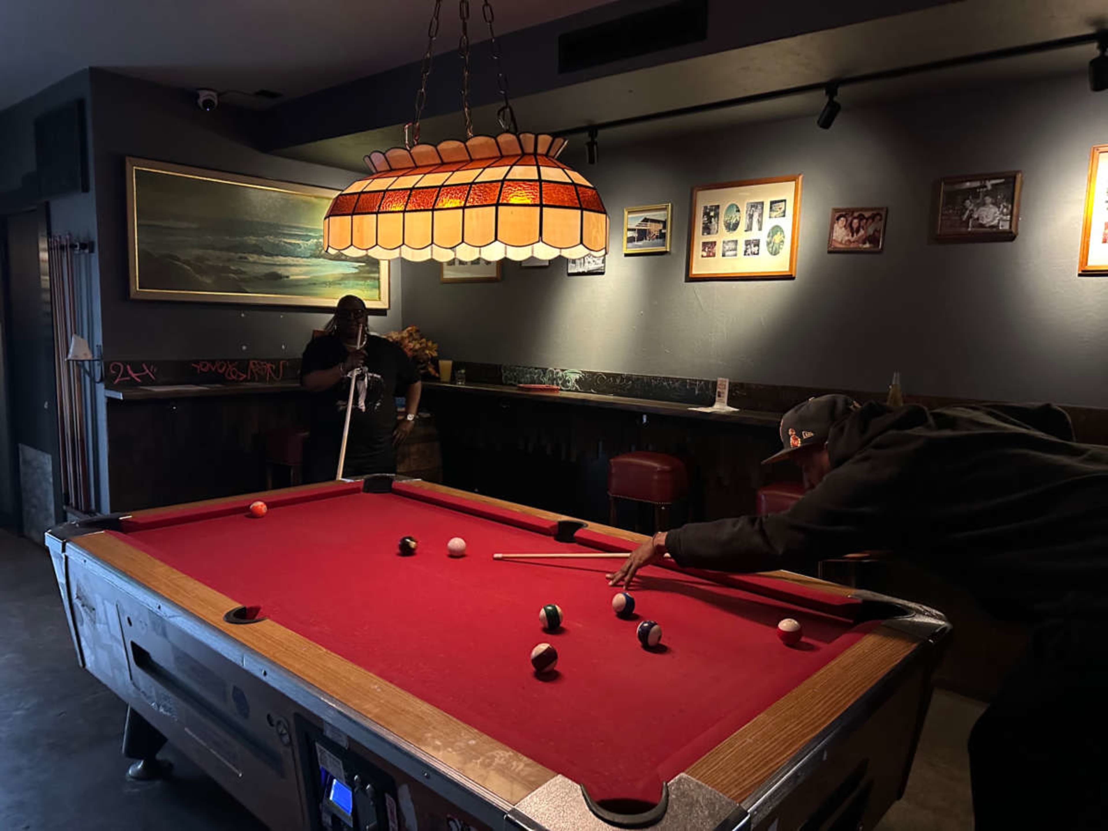 Two players are engaged in a game of pool at a well-lit table, surrounded by framed pictures and a bar in a relaxed interior setting.