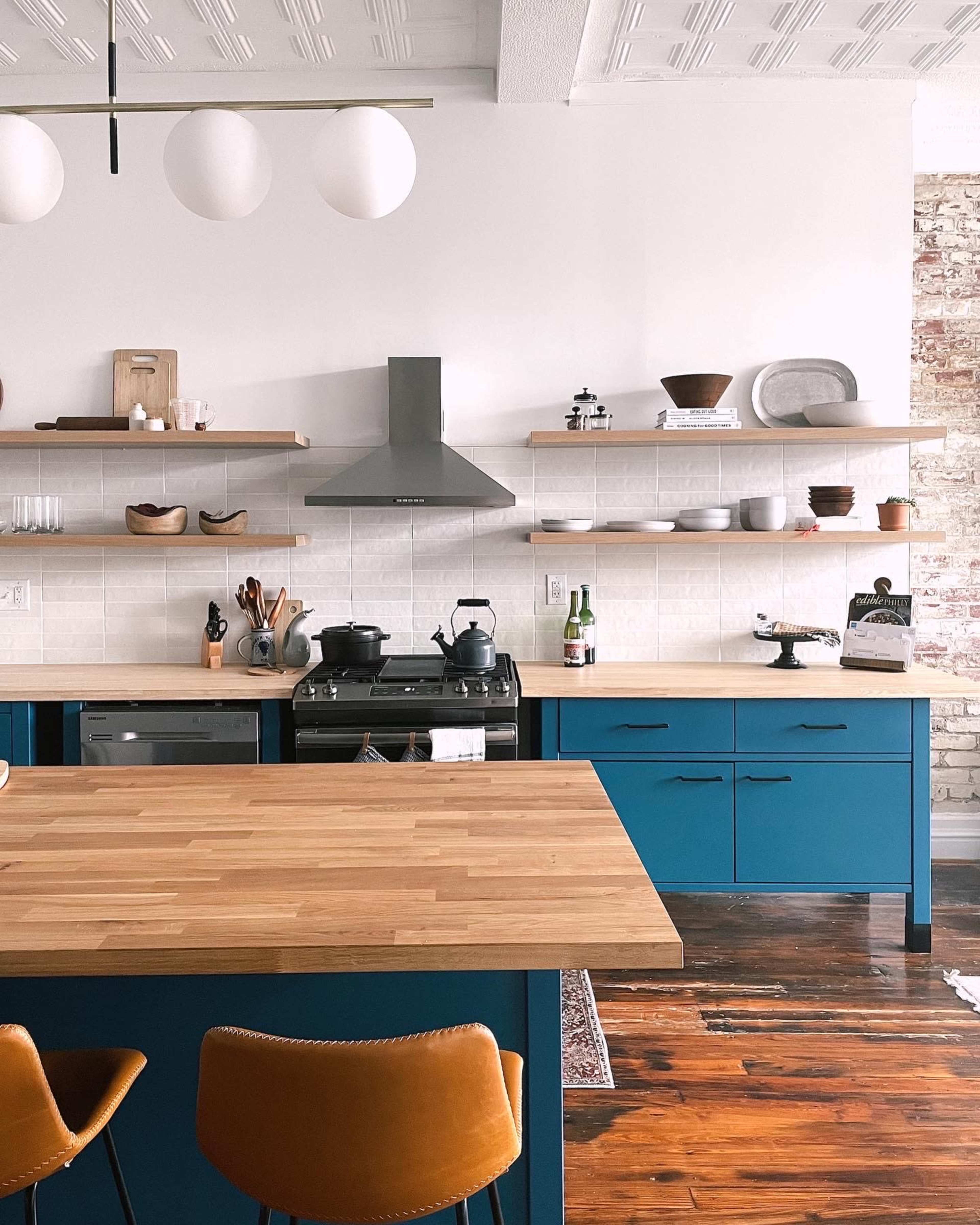 The kitchen features a modern design with teal cabinetry, wooden countertops, silver appliances, and open shelves displaying various dishes and decorations.