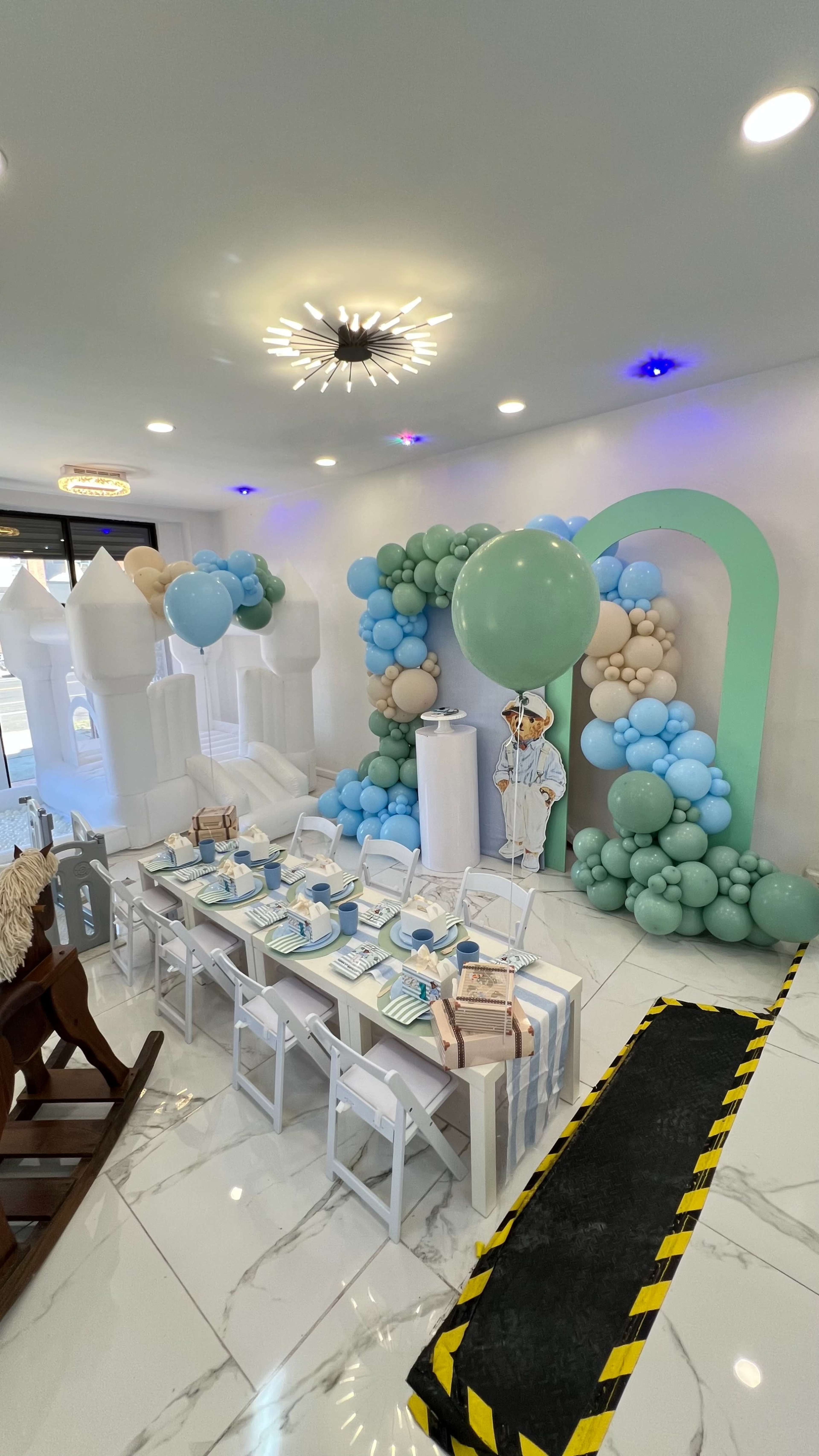 The image shows a decorated indoor space with a long dining table set for a celebration, surrounded by balloon arches in shades of blue and white, and featuring a whimsical backdrop of balloon structures.