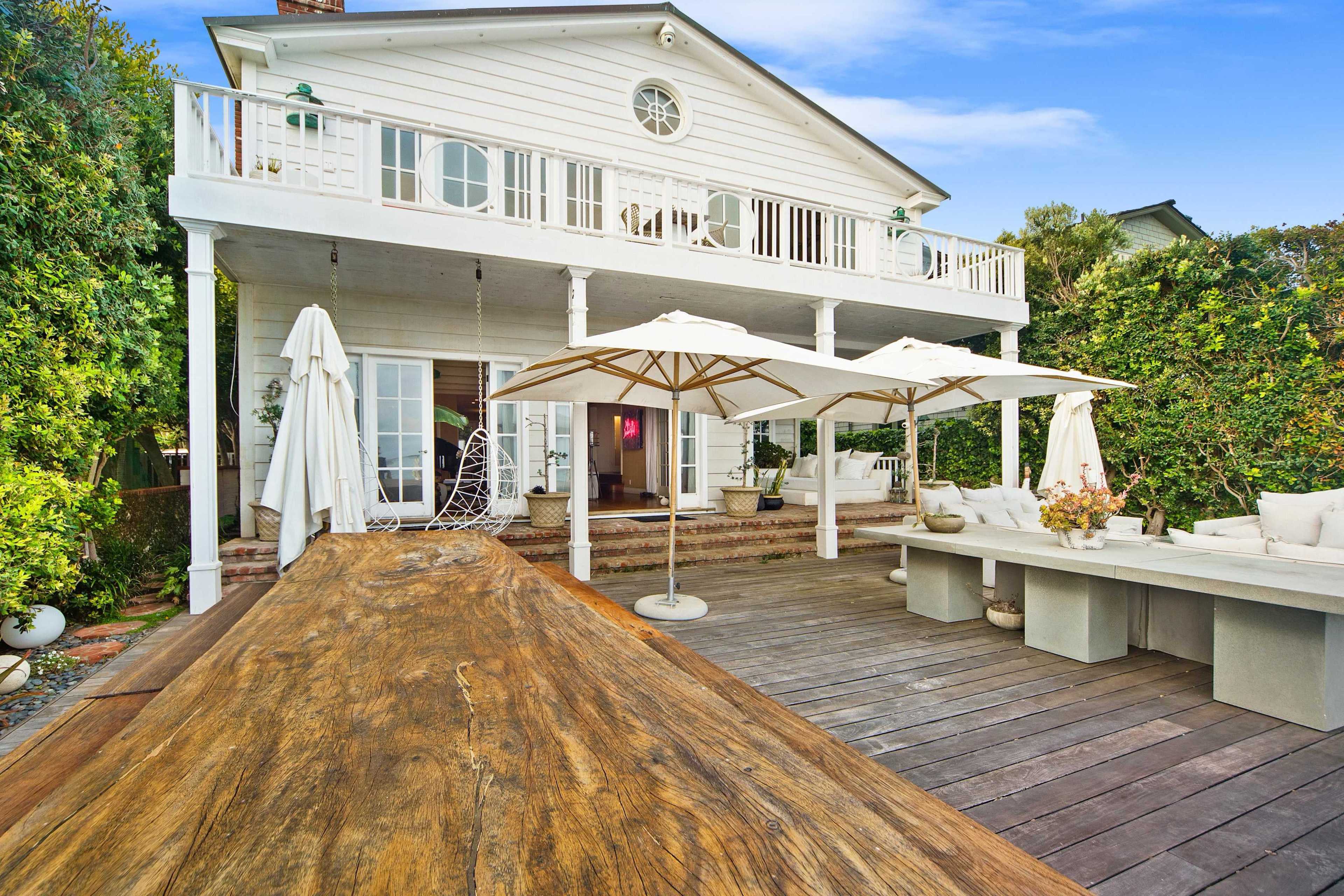 The image shows a two-story white house with a spacious wooden deck, equipped with umbrellas and a long wooden dining table, surrounded by greenery.