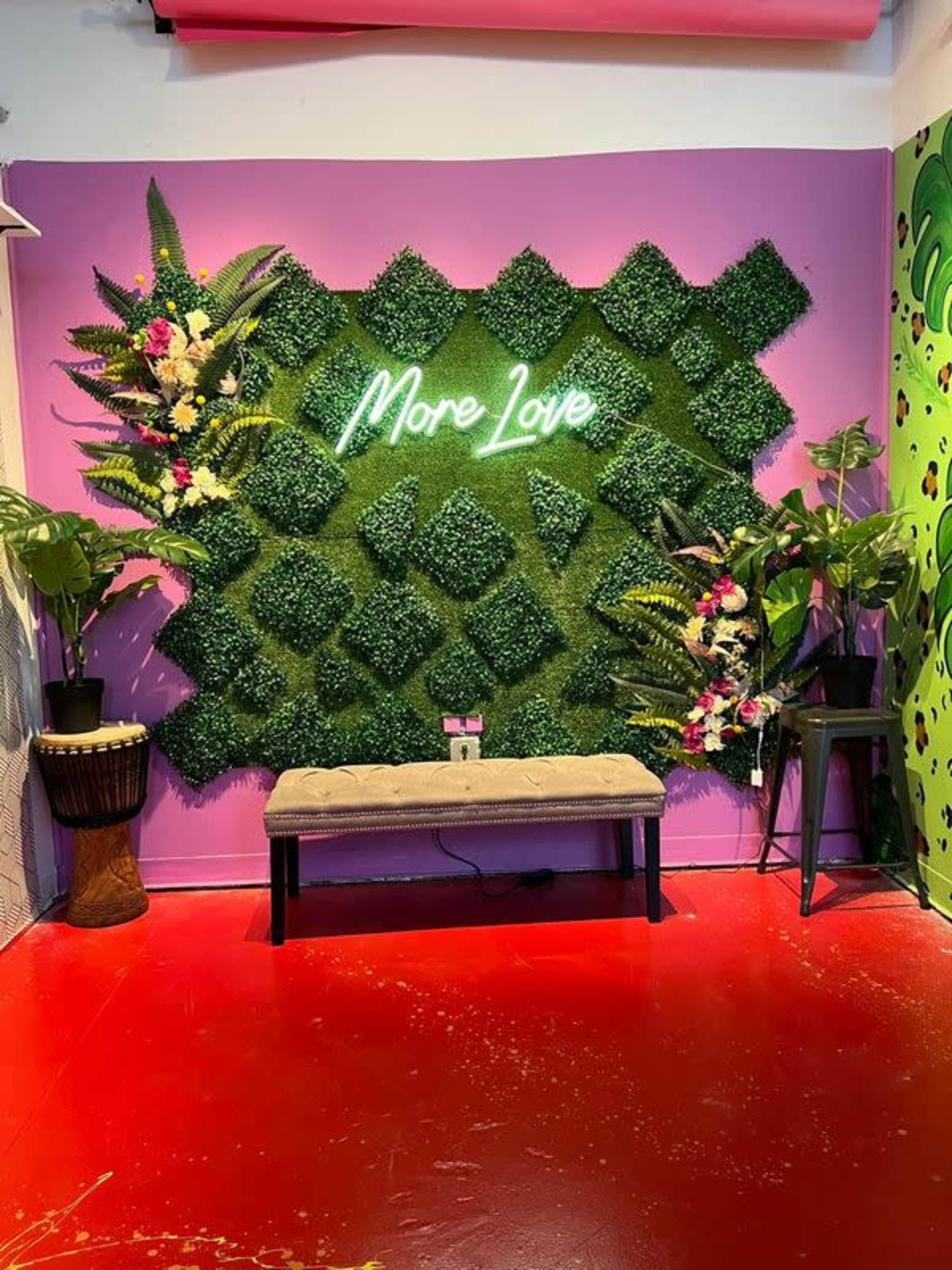 A vibrant corner of a room featuring a green artificial hedge wall with the words "More Love" in neon lights, flanked by decorative plants and a bench on a red floor.