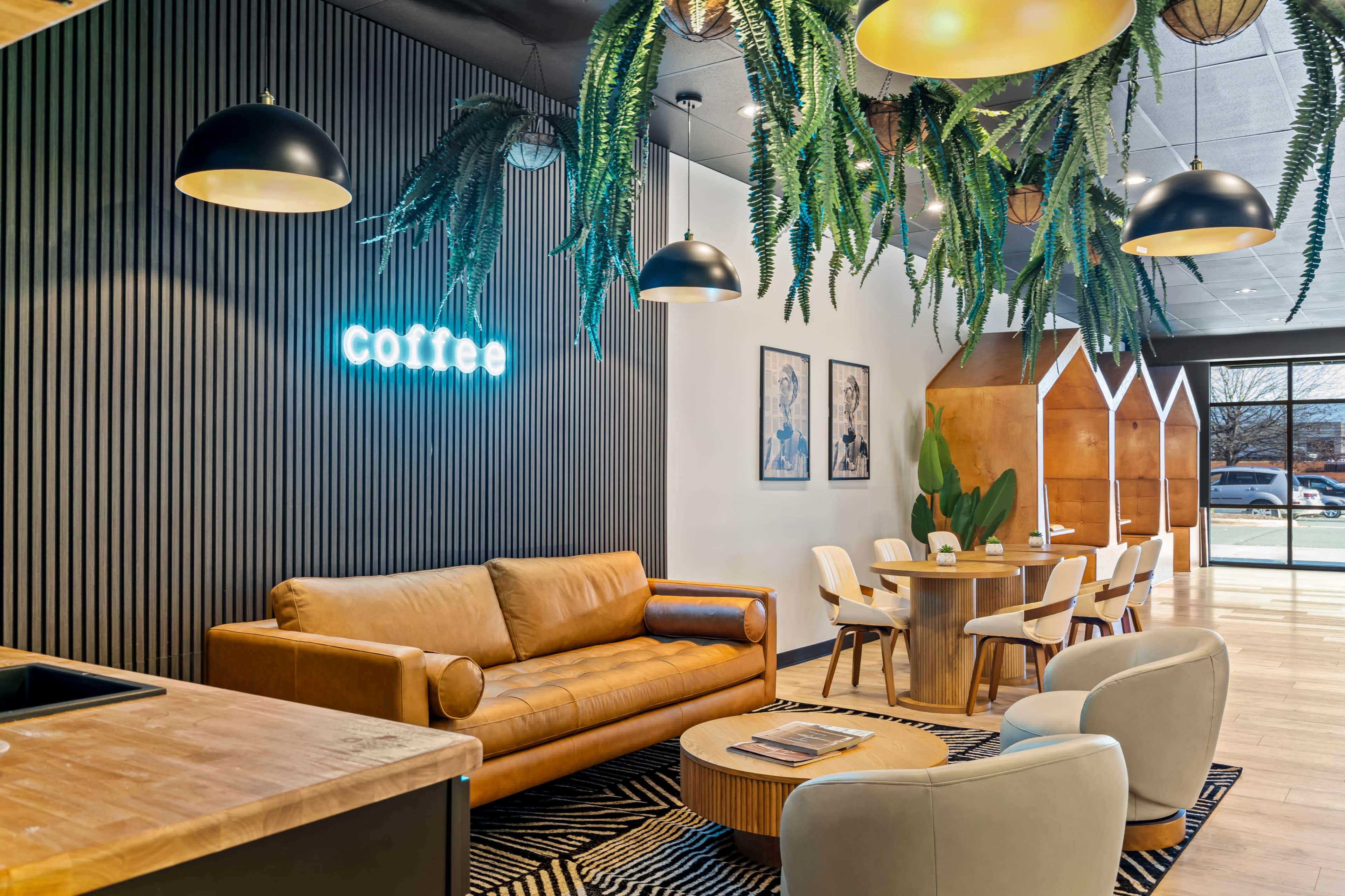 The image shows a modern coffee lounge featuring a leather sofa, a round dining table surrounded by white chairs, and hanging plants above a wooden counter.