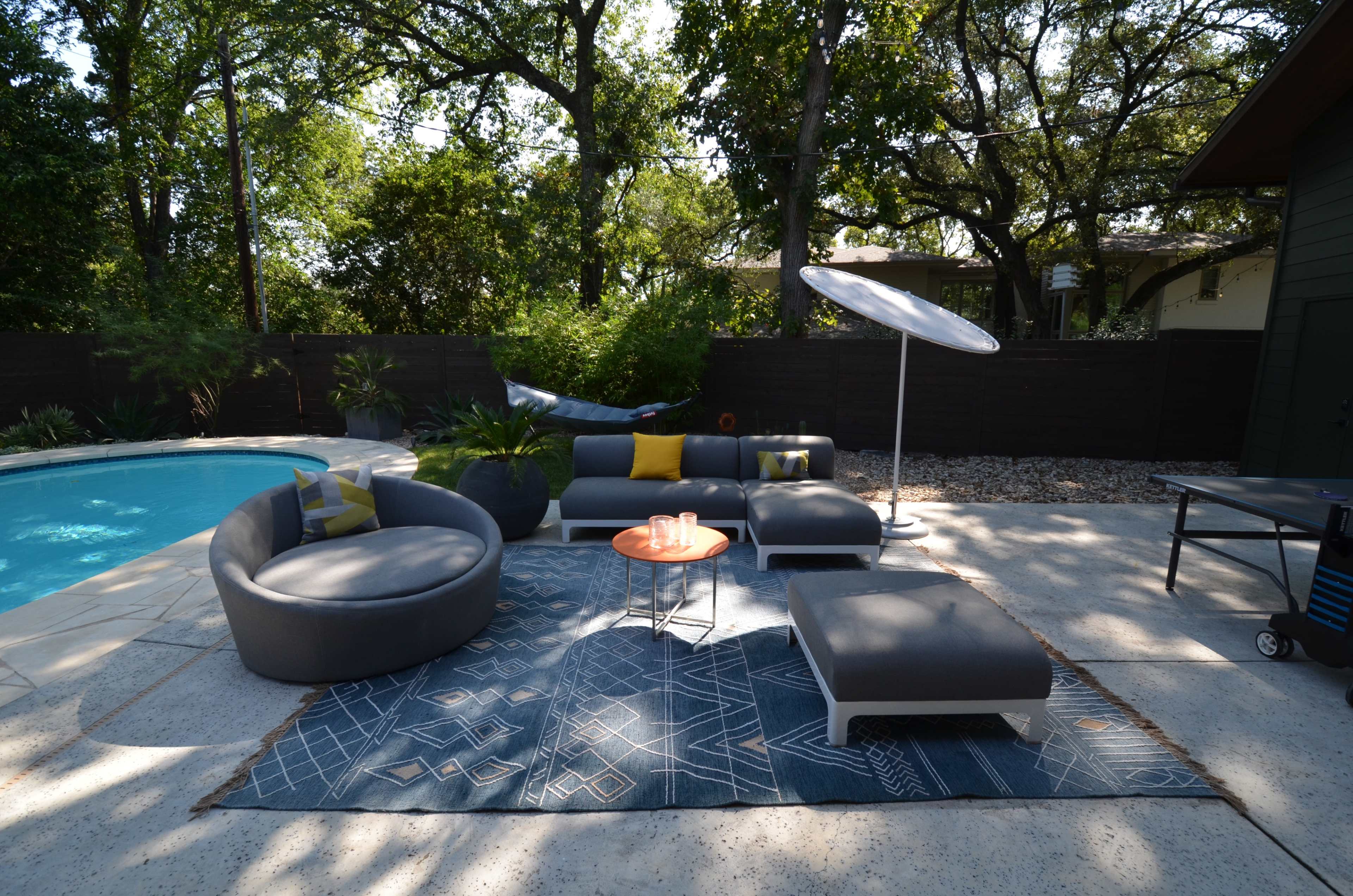 A modern outdoor seating area features circular furniture, a round table, and an umbrella beside a swimming pool, surrounded by greenery.