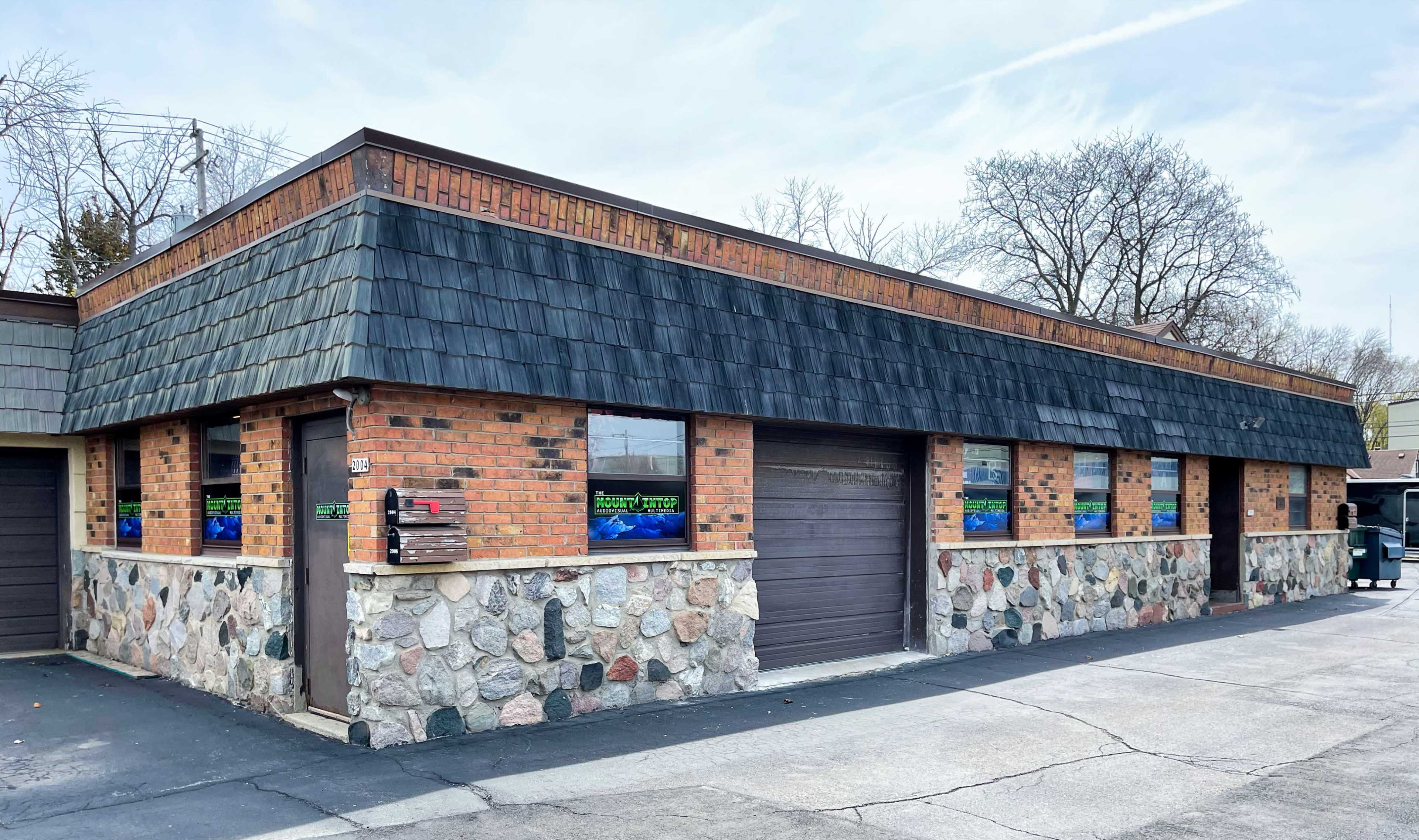 The image shows a brick building with a stone foundation, featuring large windows and a dark, shingled roof.