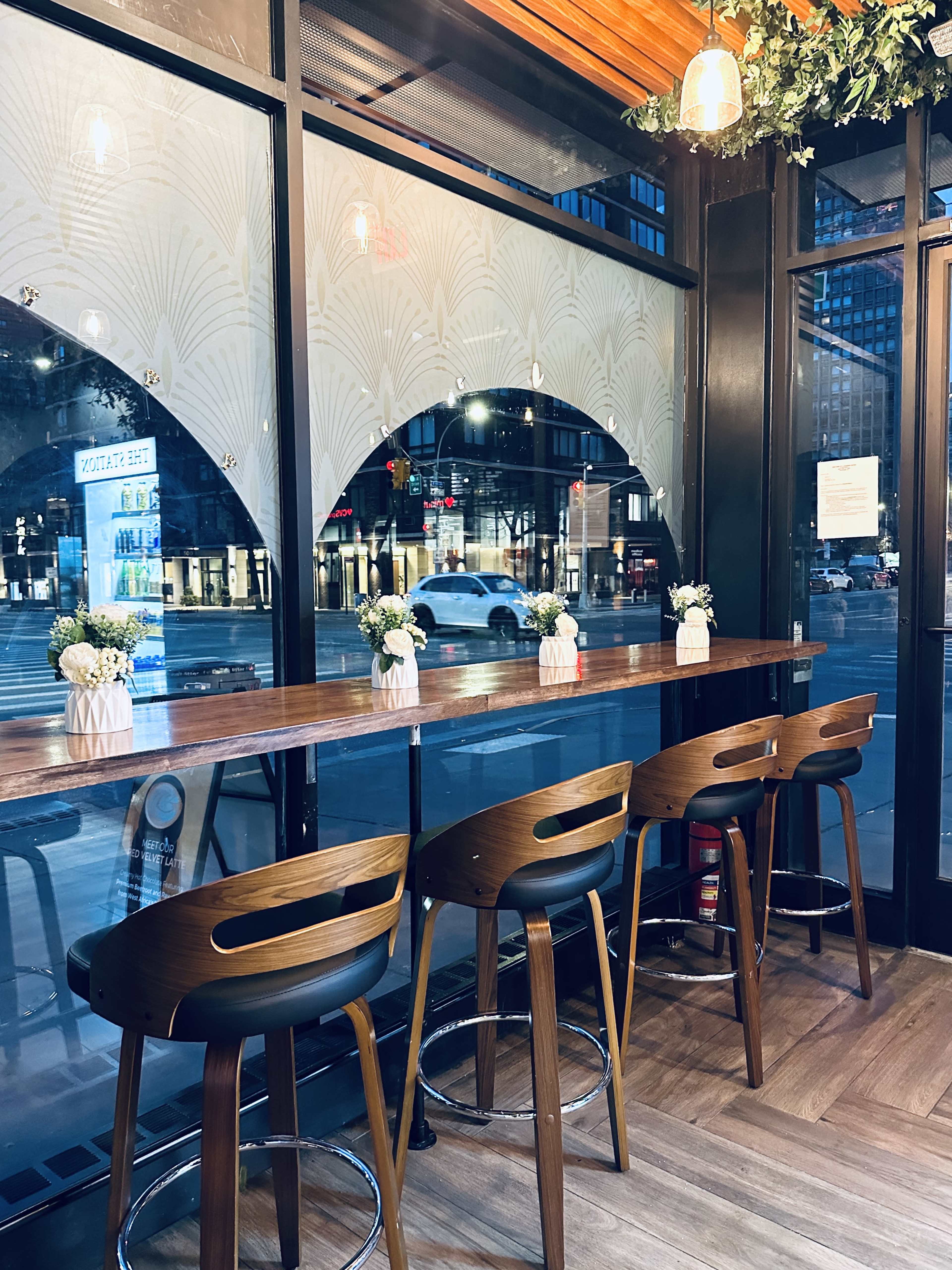 The image shows a modern cafe interior with a long wooden counter and high stools, illuminated by soft lighting, overlooking a city street through large windows.