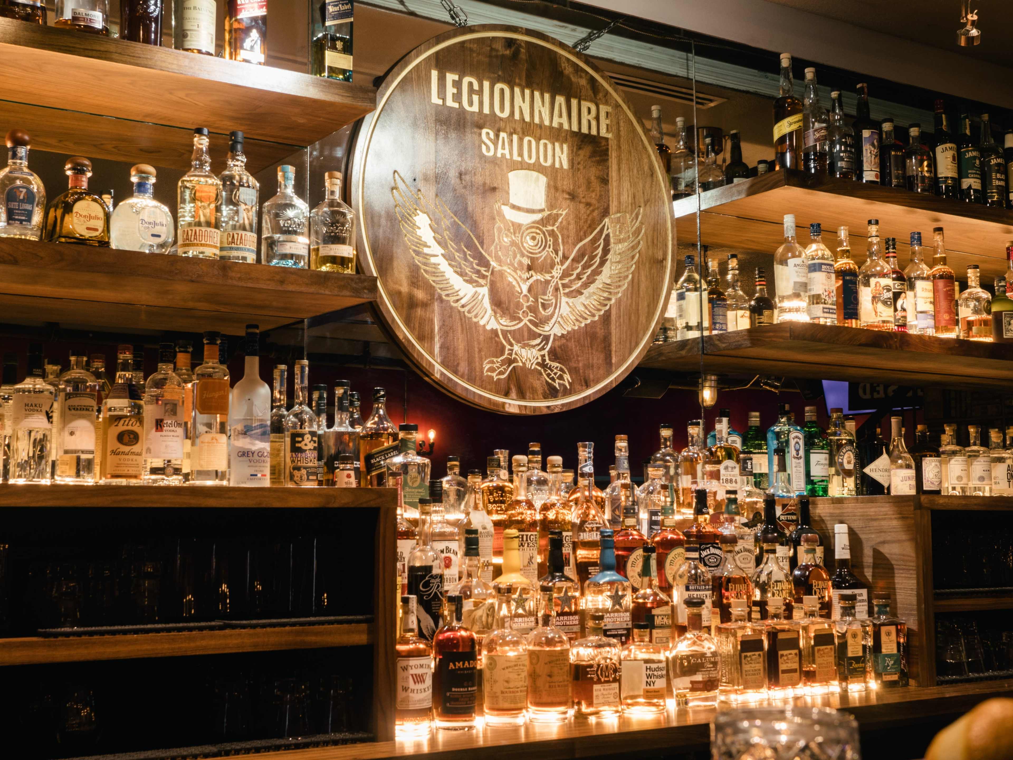 The image shows a well-stocked bar featuring numerous bottles of liquor displayed on wooden shelves beneath a round sign labeled "Legionnaire Saloon."