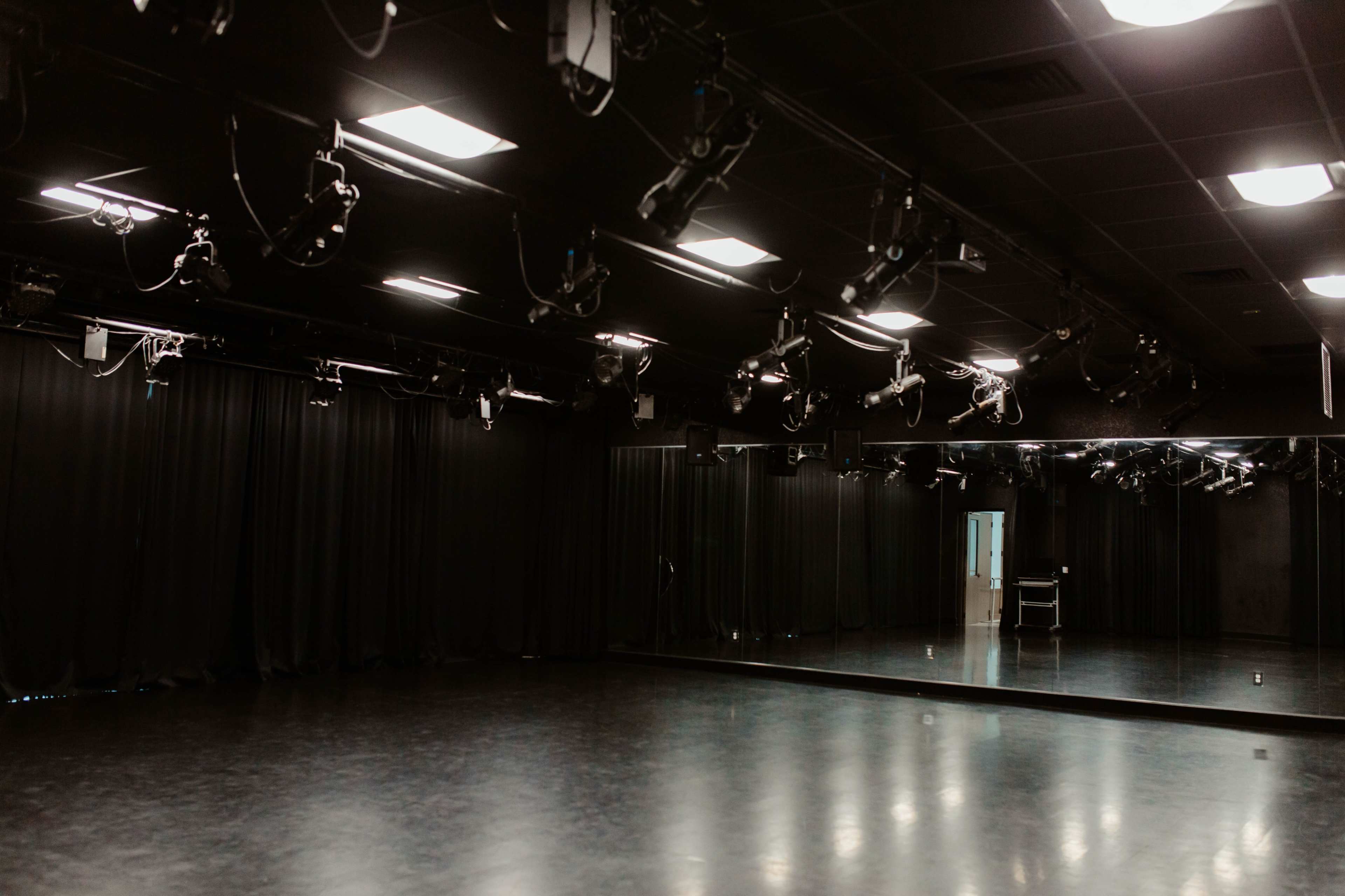 The image shows a dimly lit rehearsal space with black curtains, multiple stage lights, and a mirrored wall.