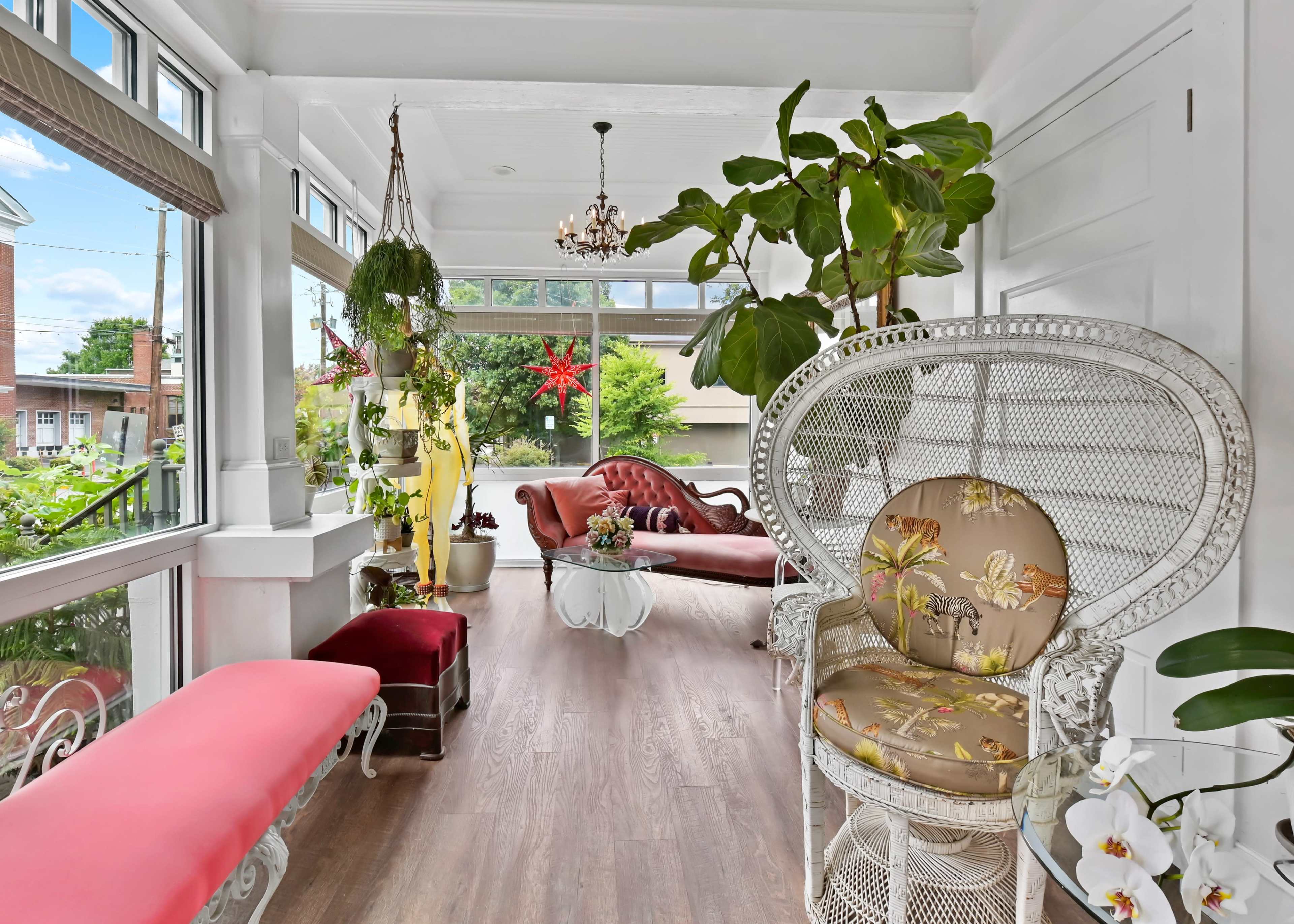 A bright sunroom with a wicker chair, a red couch, and various plants surrounding a wooden floor.