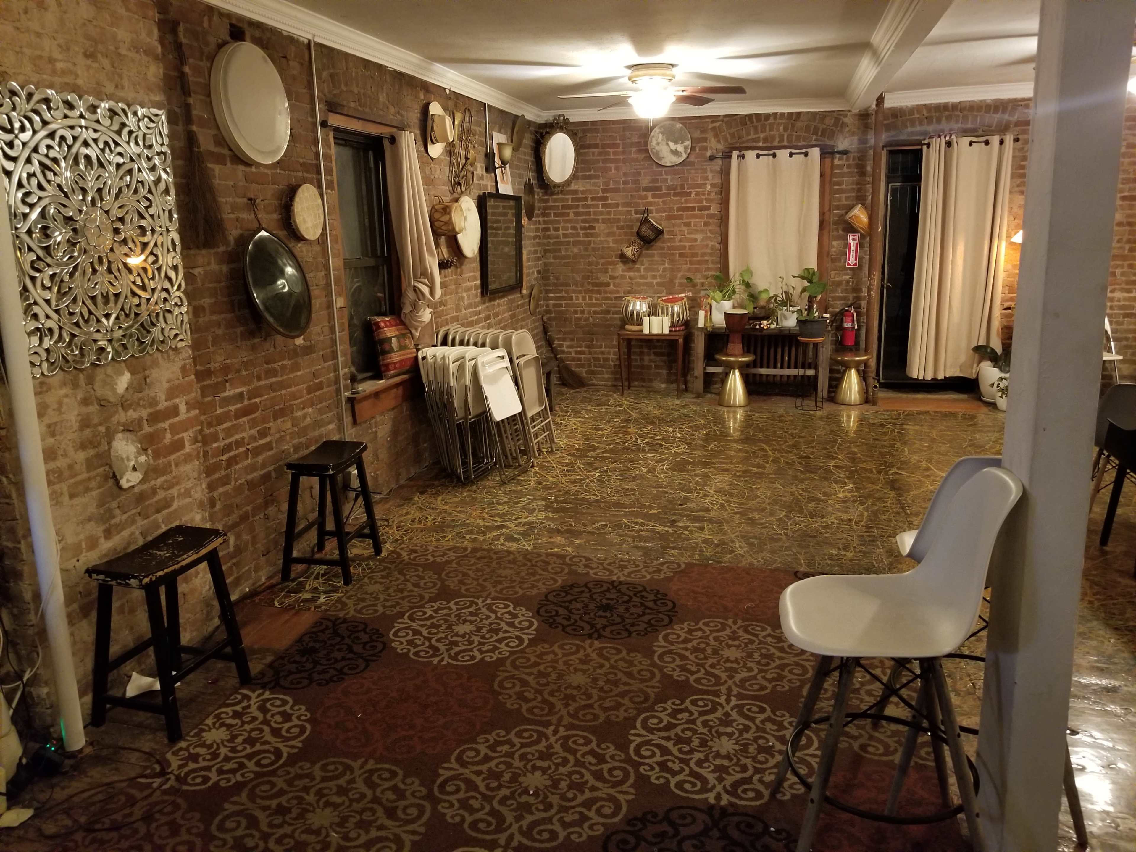 A room with exposed brick walls, a patterned floor, and various pieces of furniture, including stools and tables, arranged in a sparse layout.