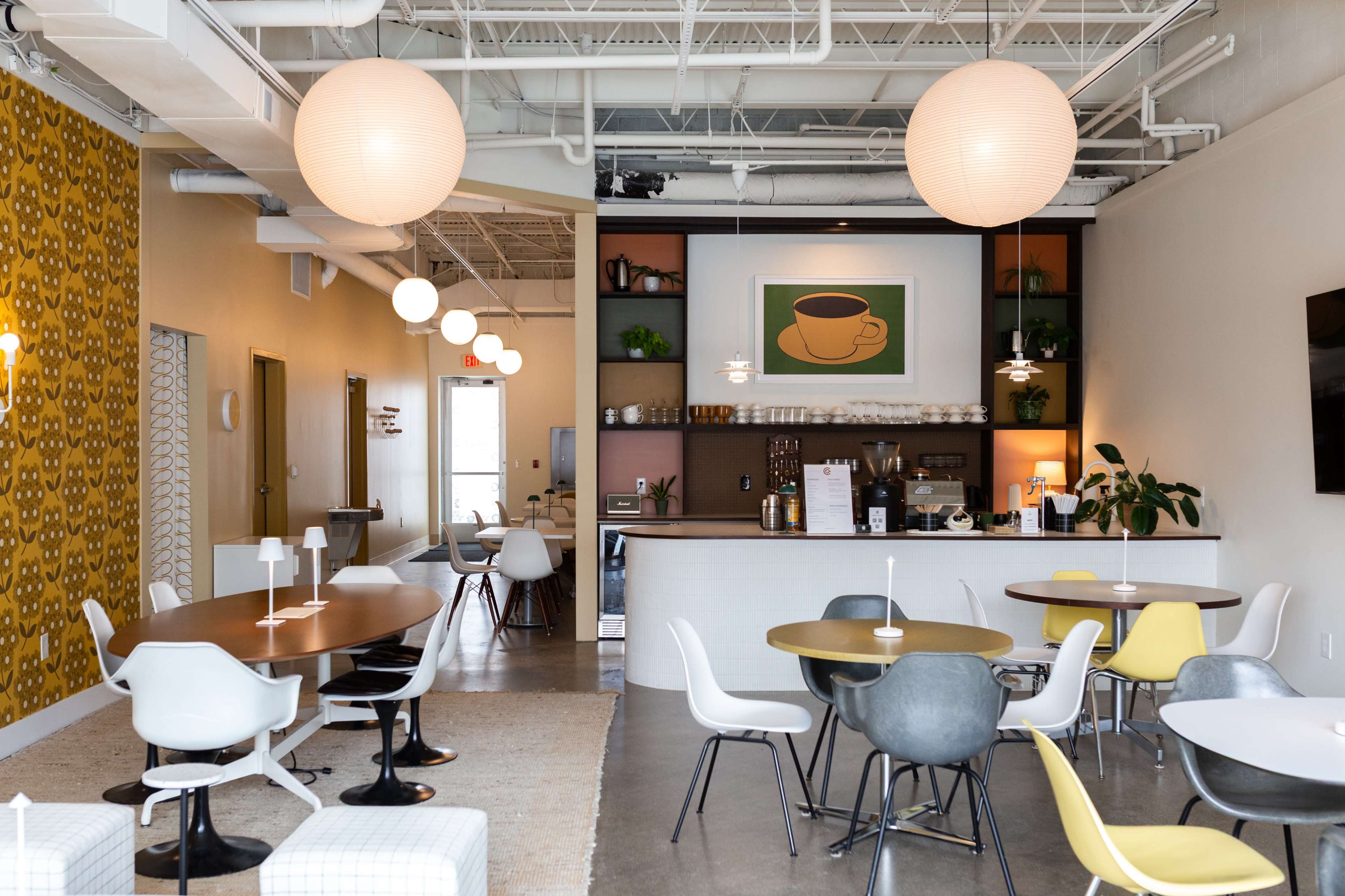 The image shows a modern coffee shop interior with a combination of round tables and chairs, a coffee bar in the background, and decorative lighting.