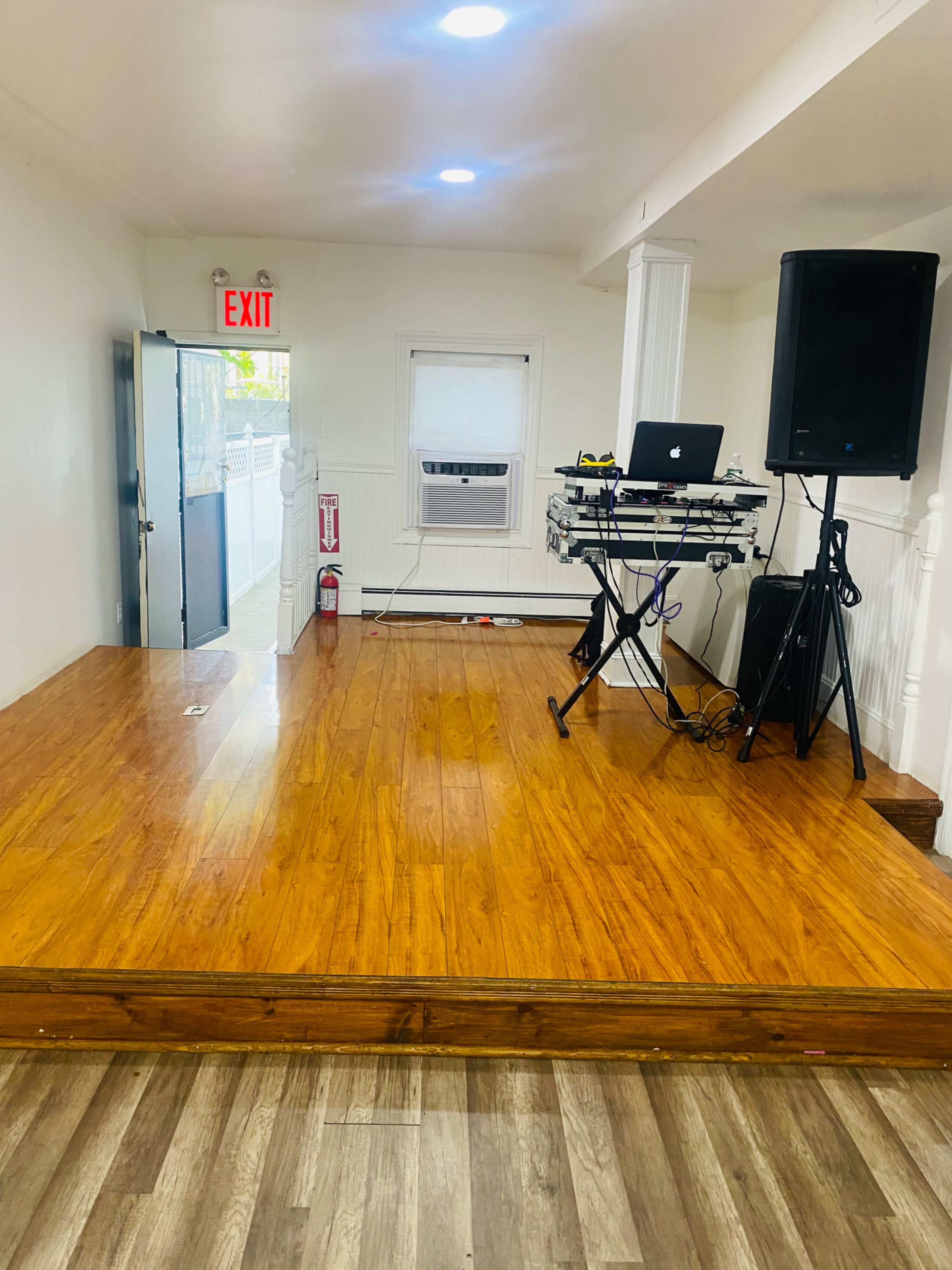 A spacious room with wooden floors, an exit sign, and a DJ setup including a mixer and speakers.