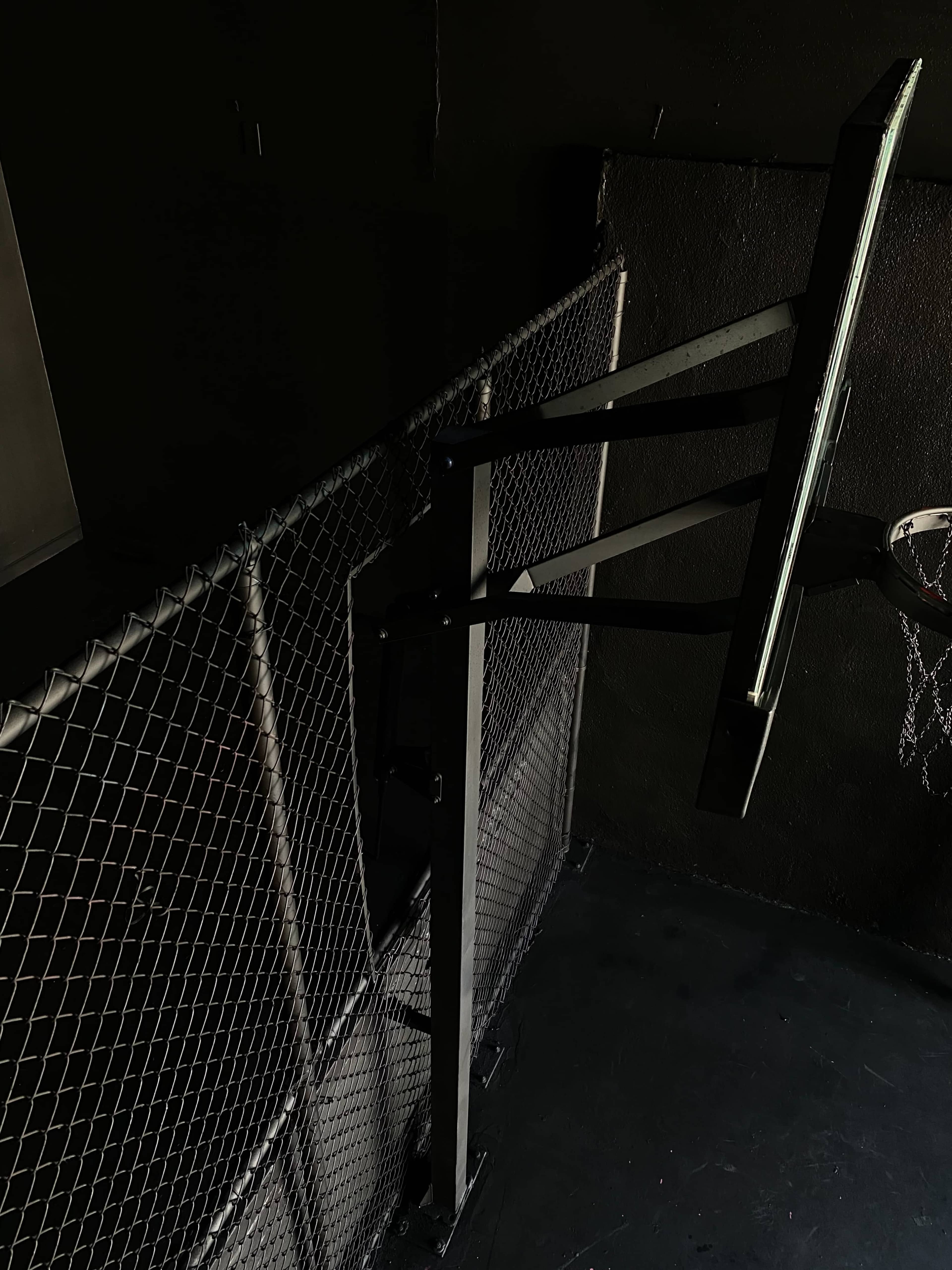 A basketball hoop mounted on a wall next to a chain-link fence in a dimly lit area.